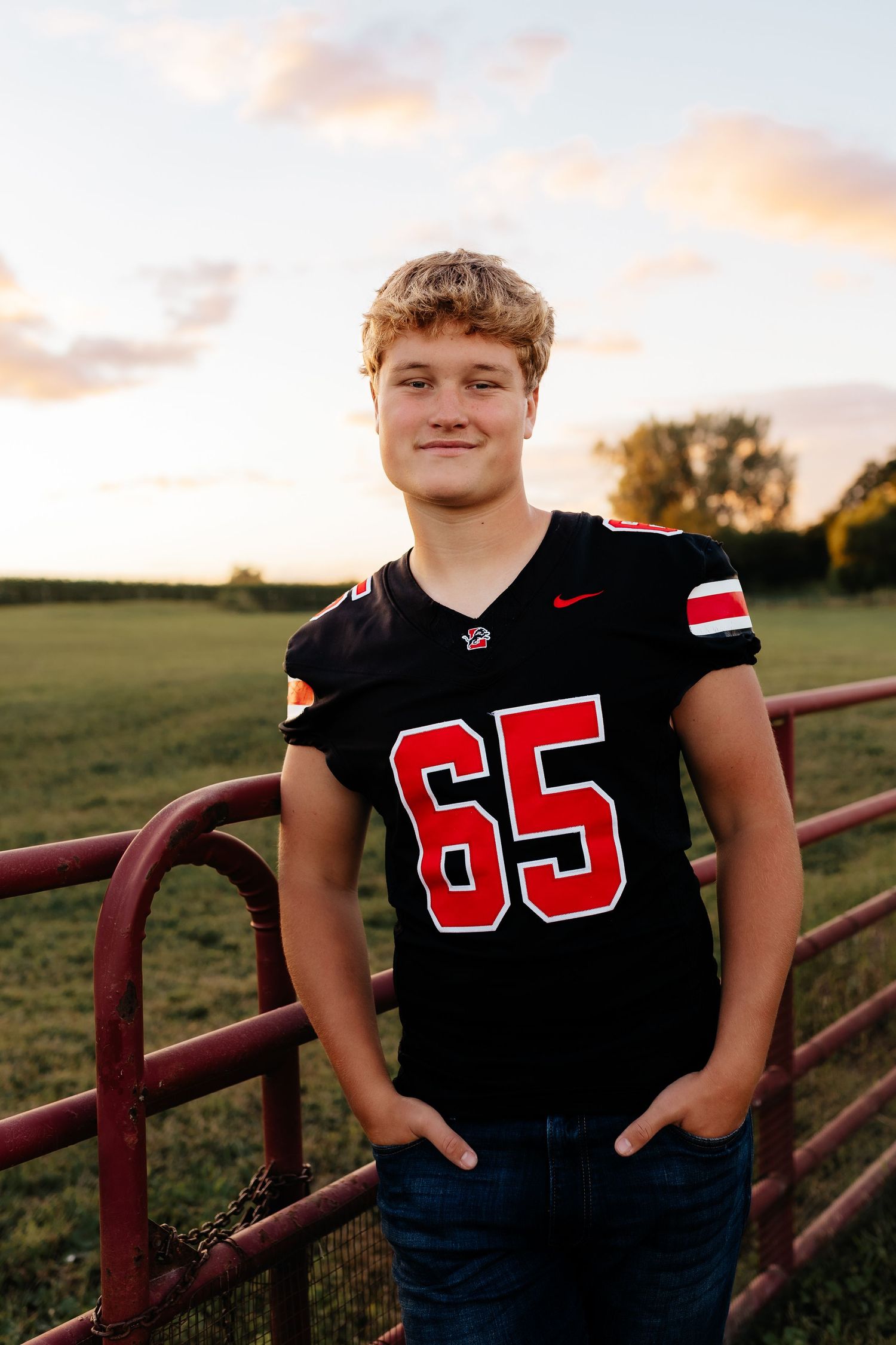 Teen boy in black number 65 football jersey smiling at sunset by a red farm gate in a green field.