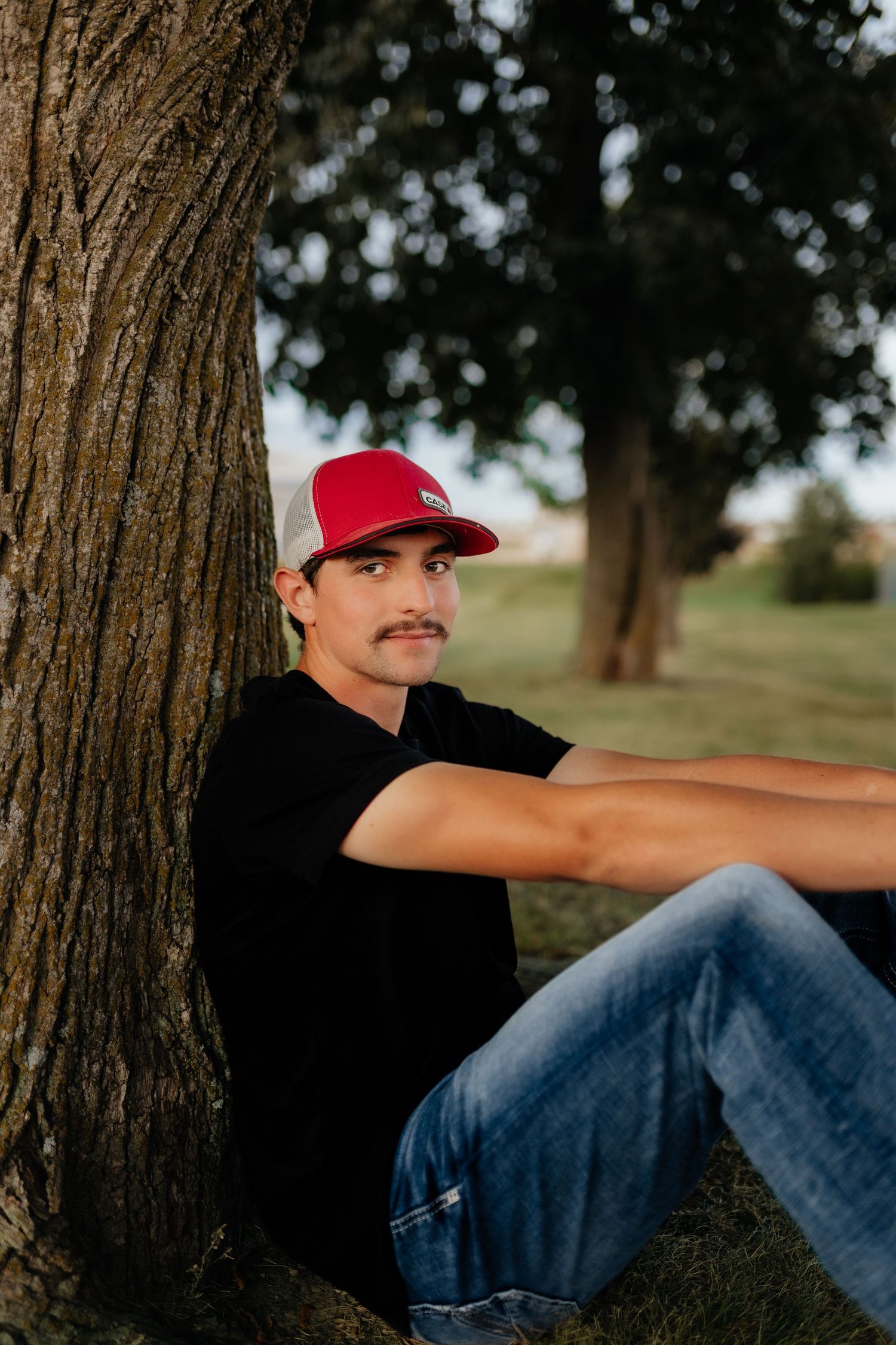 A young man with a mustache wearing a red cap and black shirt leans against a tree outdoors.