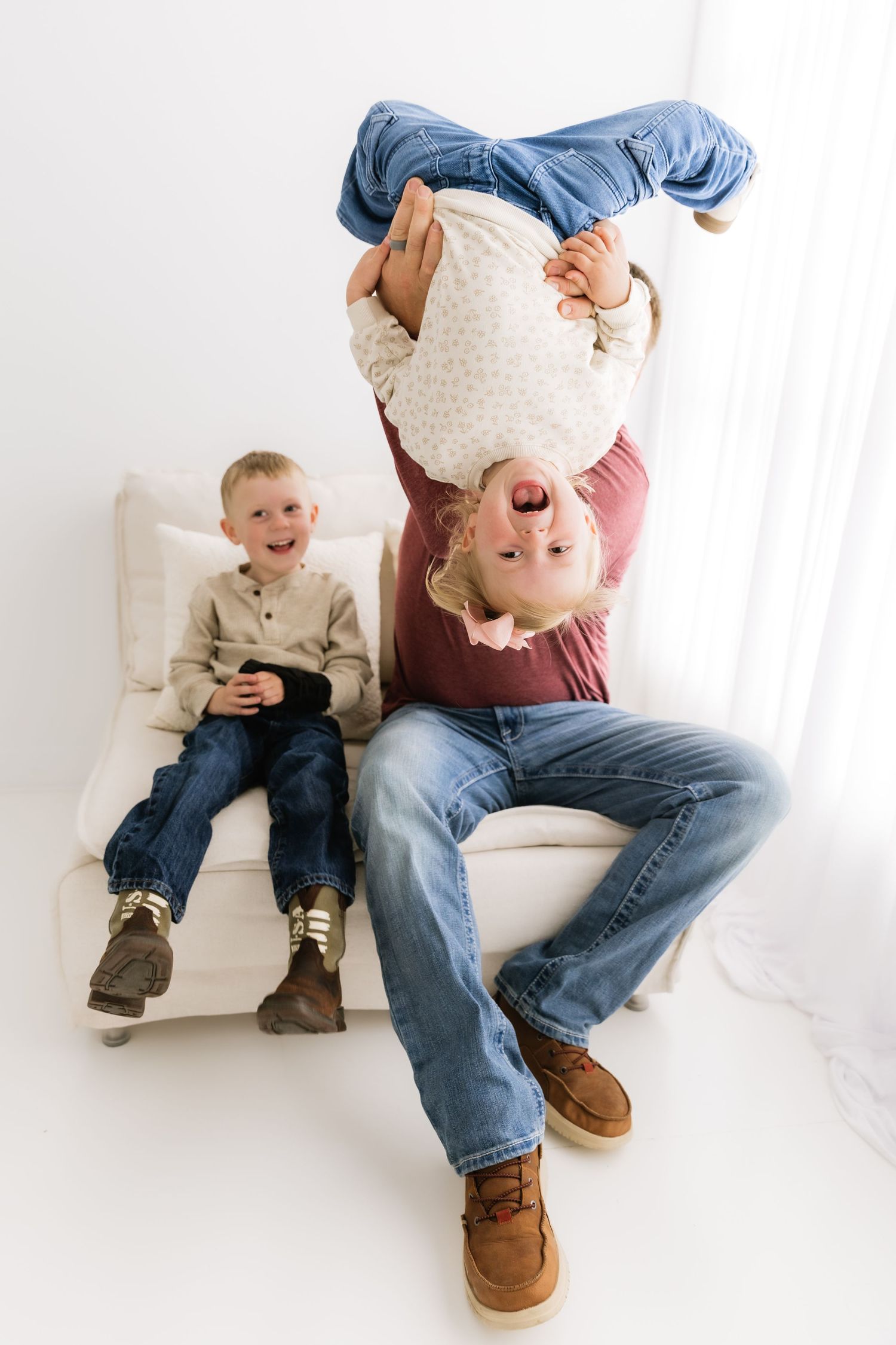Father holds laughing child upside down while son watches and smiles on white studio bench.