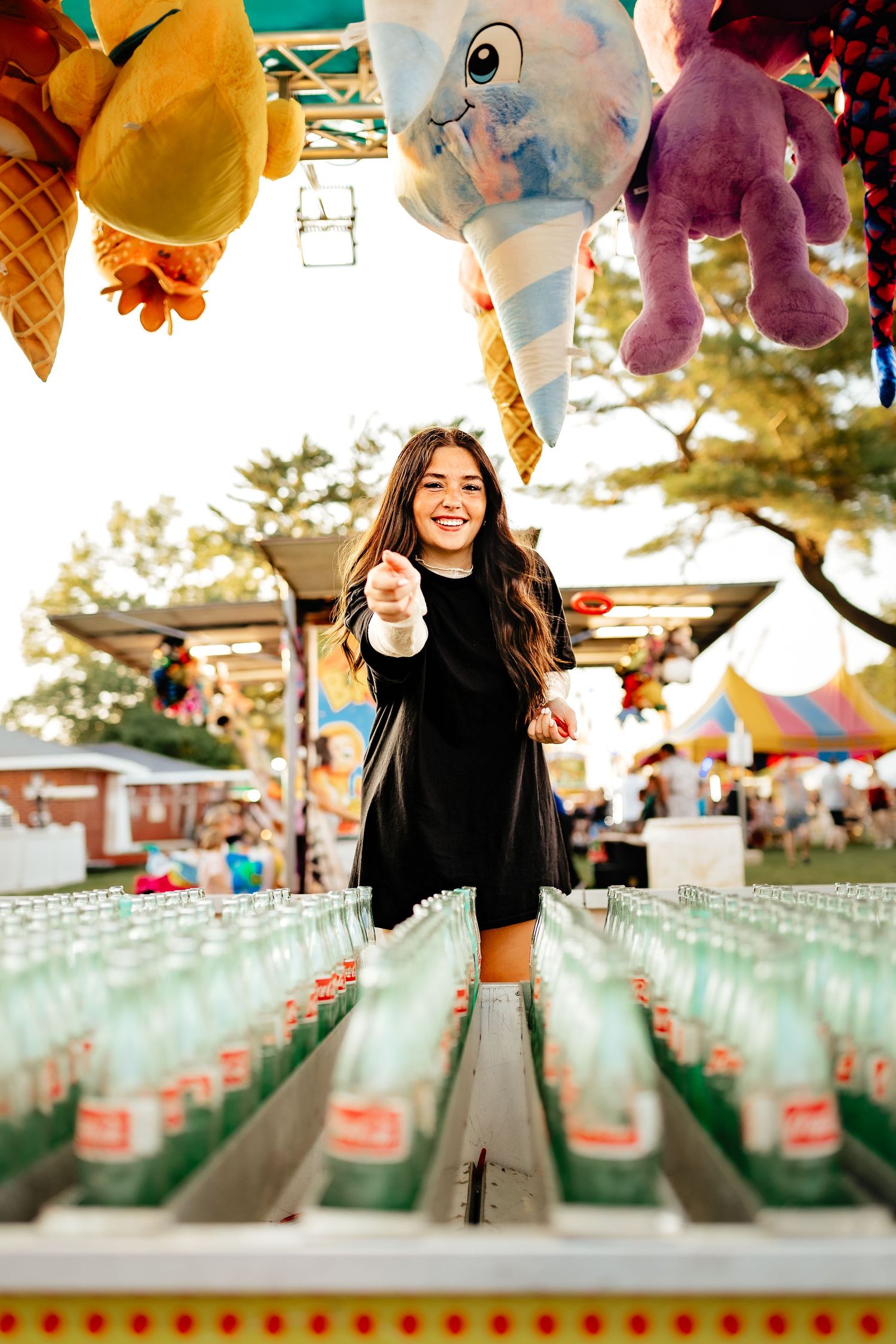Smiling young woman in black dress reaching toward camera at a carnival bottle ring toss game.