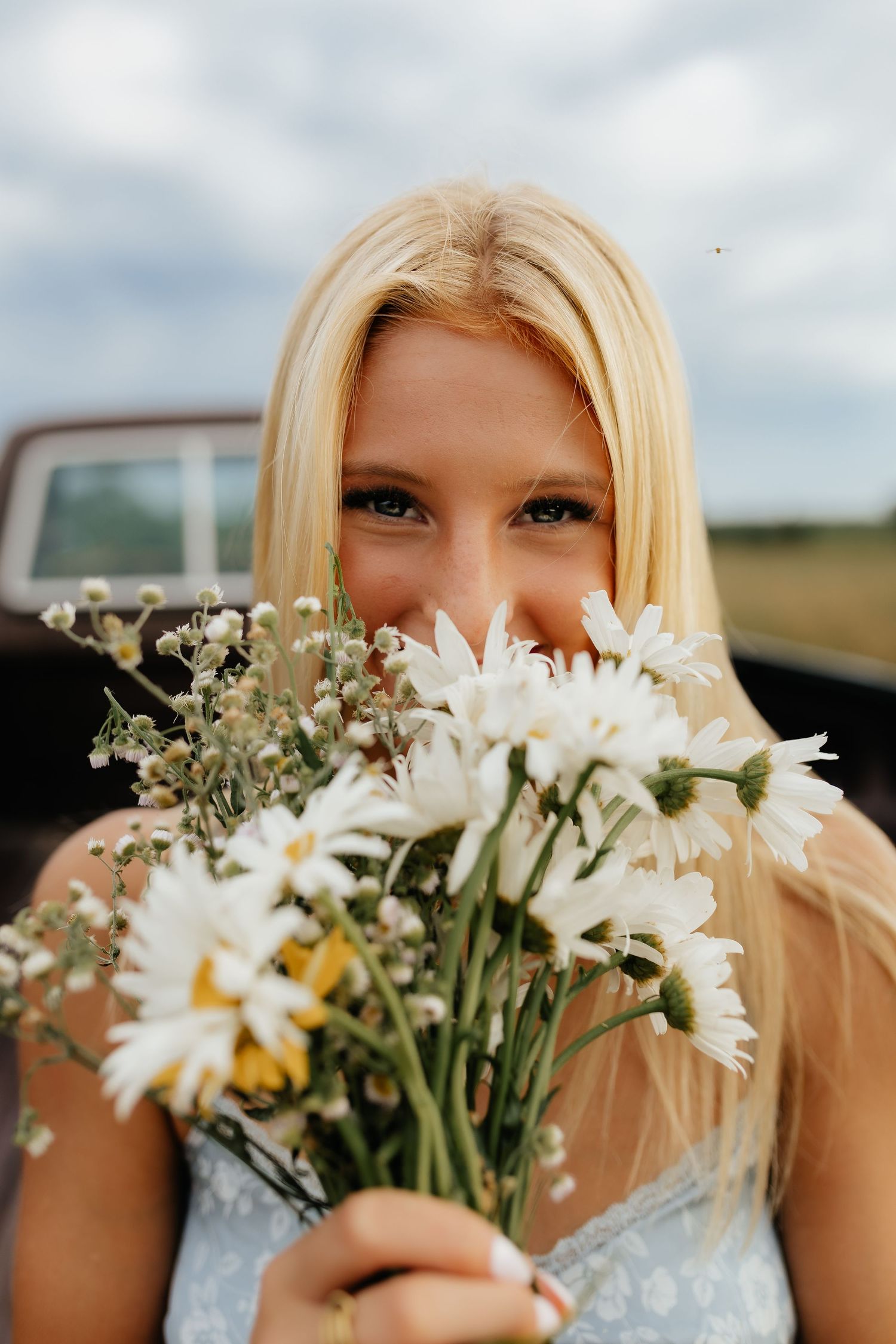 A blonde girl holds a bouquet of white daisies up to her face outdoors near a vintage truck.
