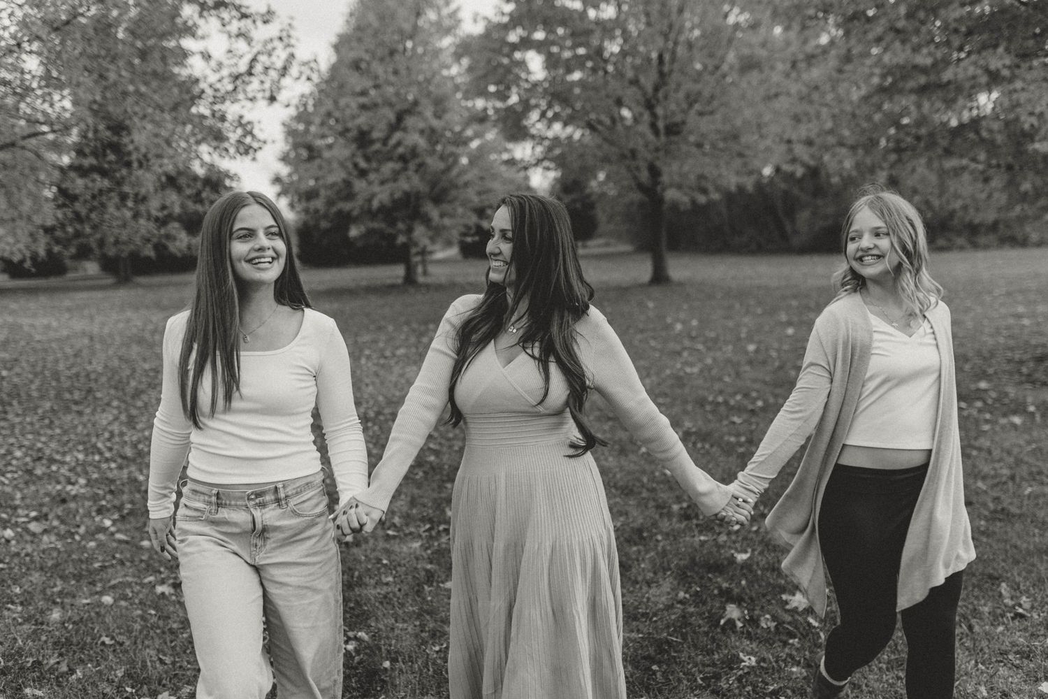 Black and white photo of three women holding hands and walking together in a park during fall season.