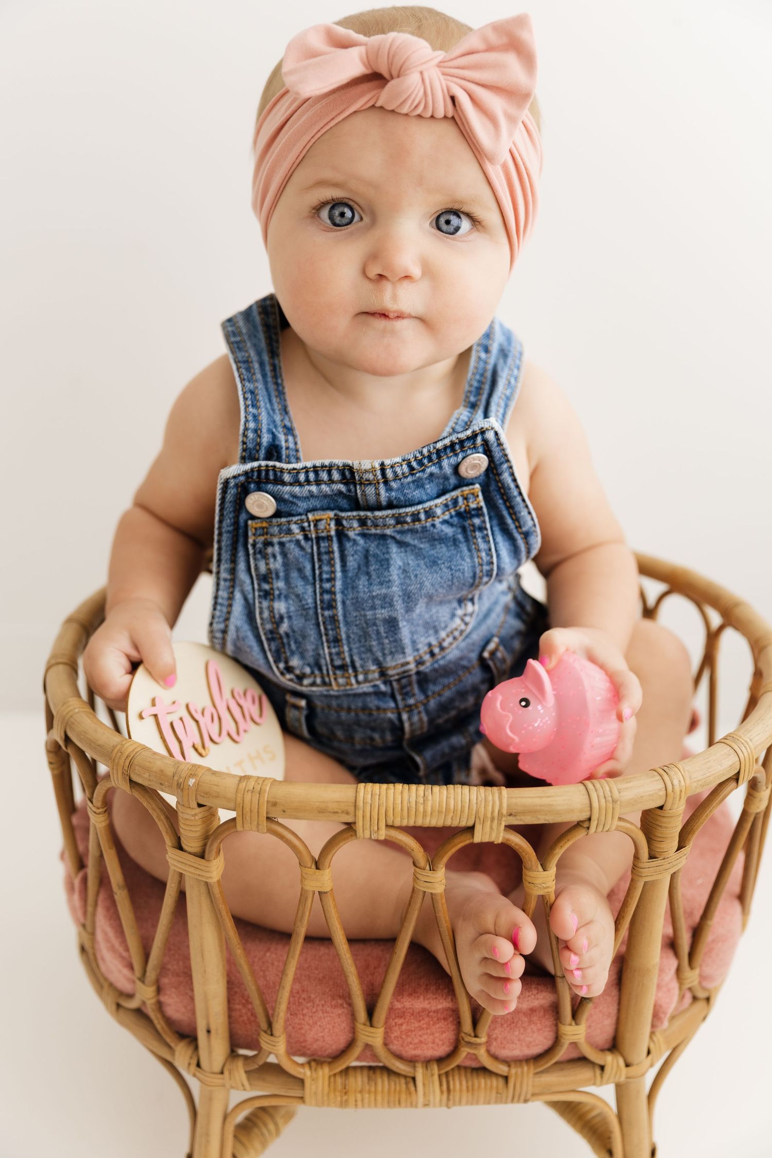 Baby girl in denim overalls and pink bow headband sits in rattan basket holding piggy bank.