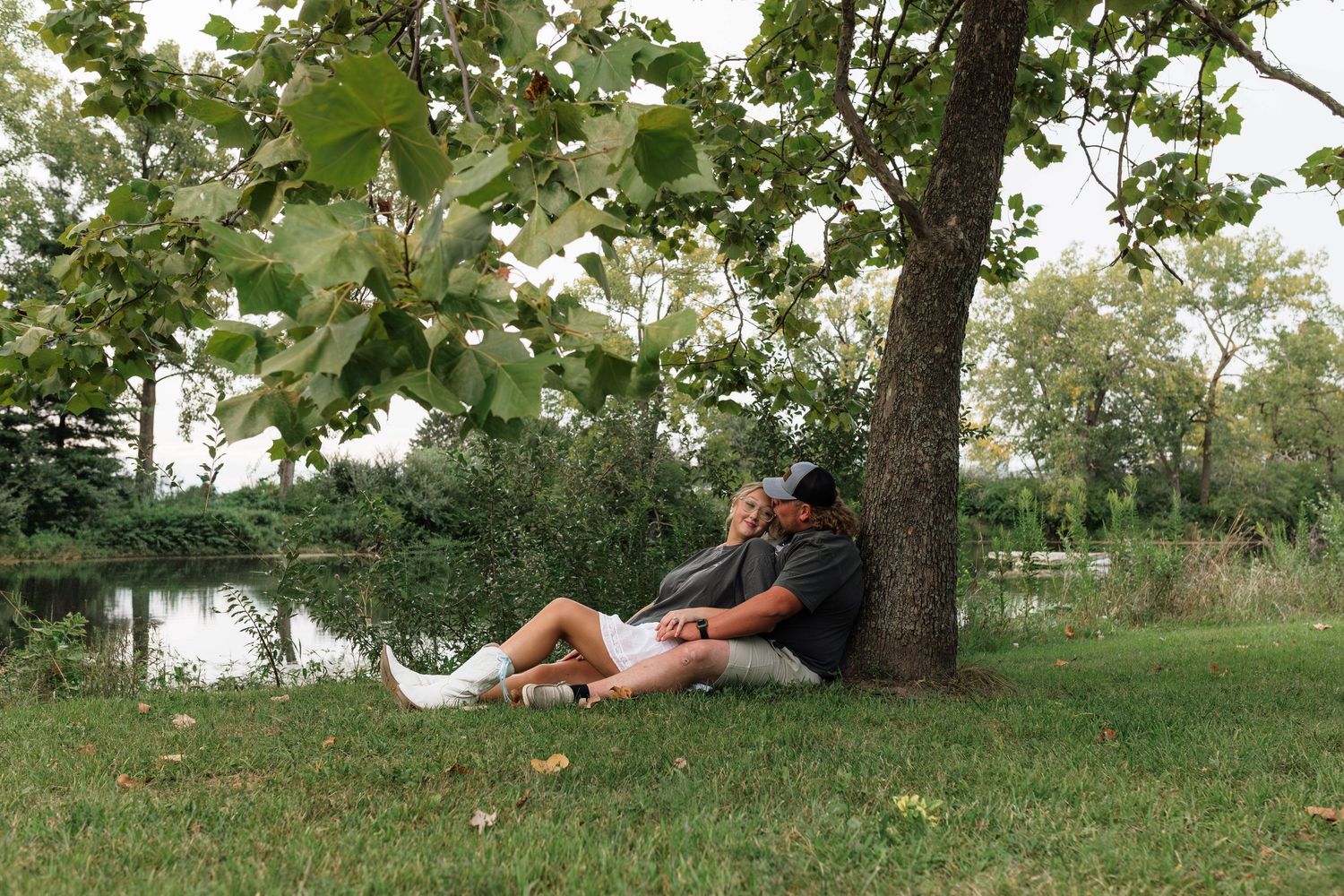 Couple sitting together leaning against a tree by a serene lakeside setting.