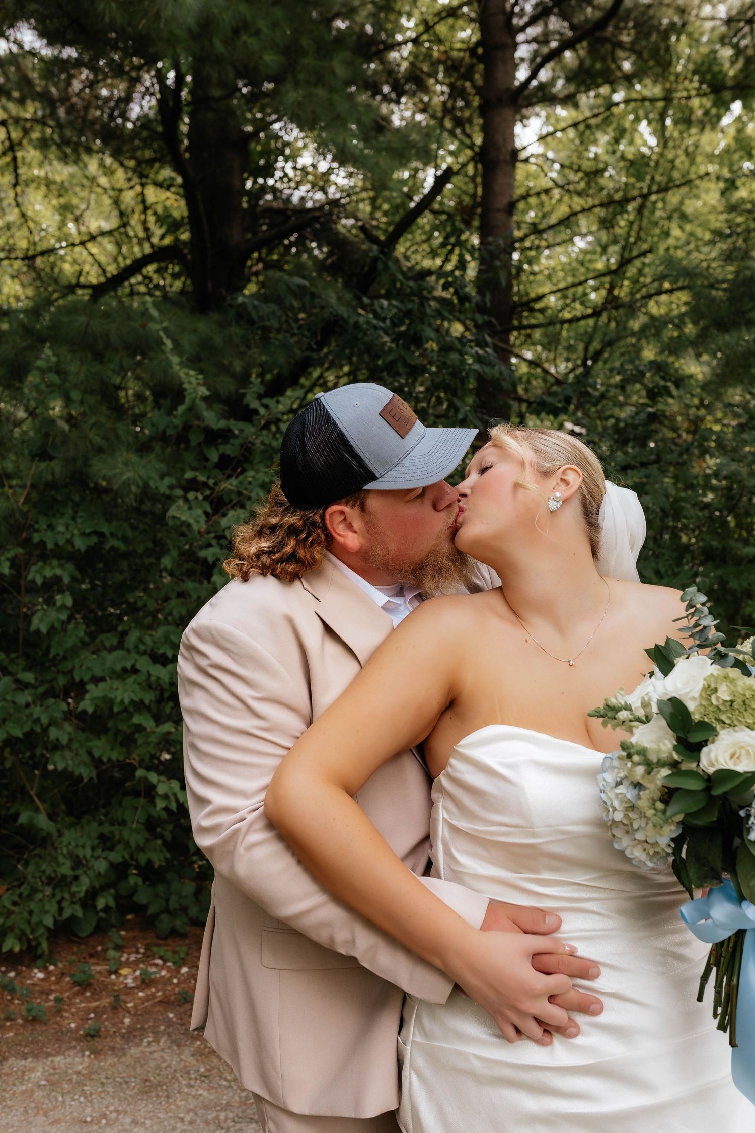 Newlyweds share romantic kiss outdoors, groom in beige suit and cap, bride in strapless white gown.