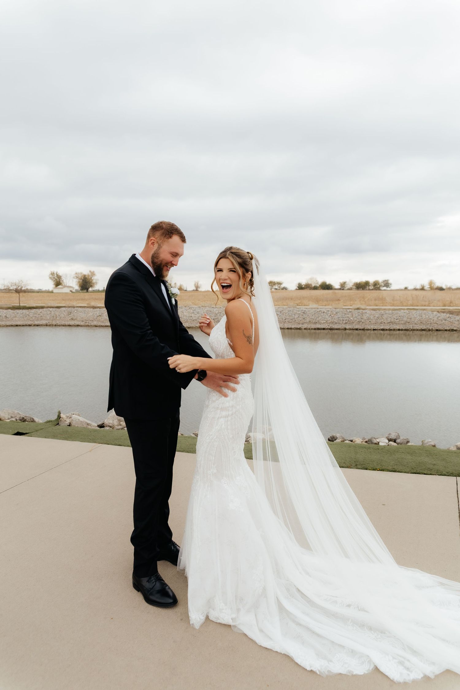 Laughing bride in lace mermaid gown with cathedral veil holds grooms hands during first look by lake.