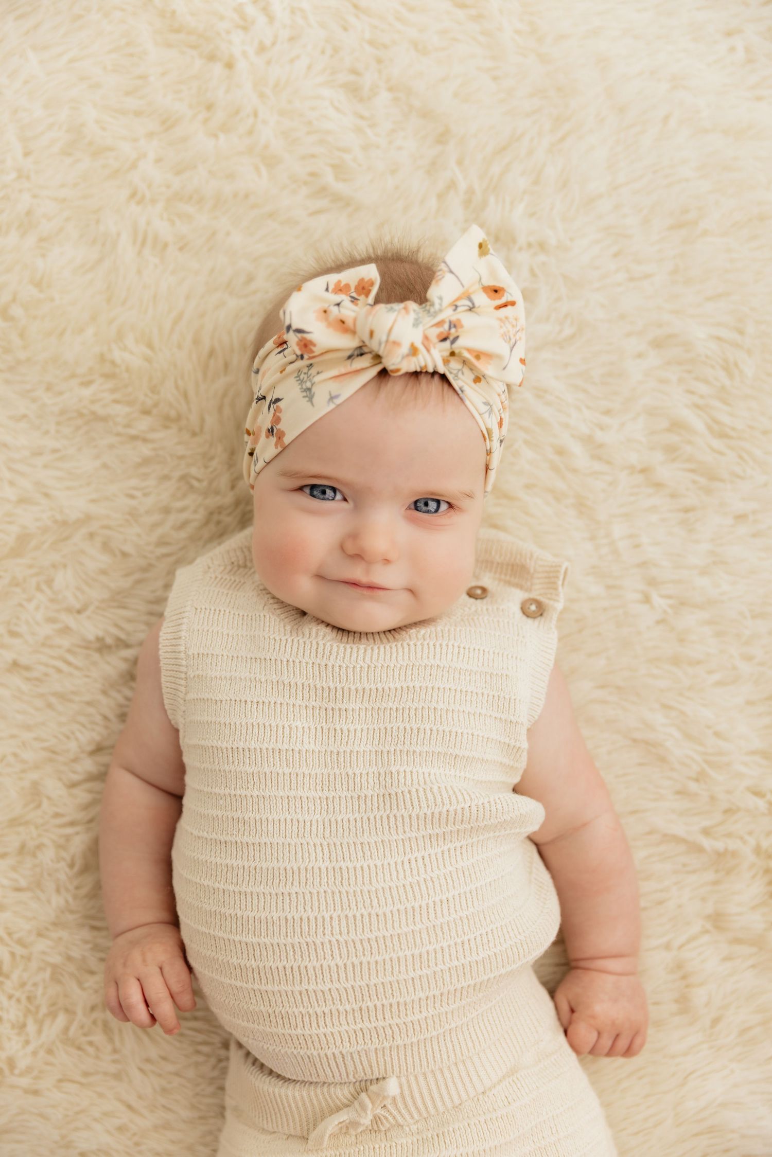 Smiling baby girl with blue eyes wearing a cream knit romper and floral bow headband lying on a fluffy white blanket.