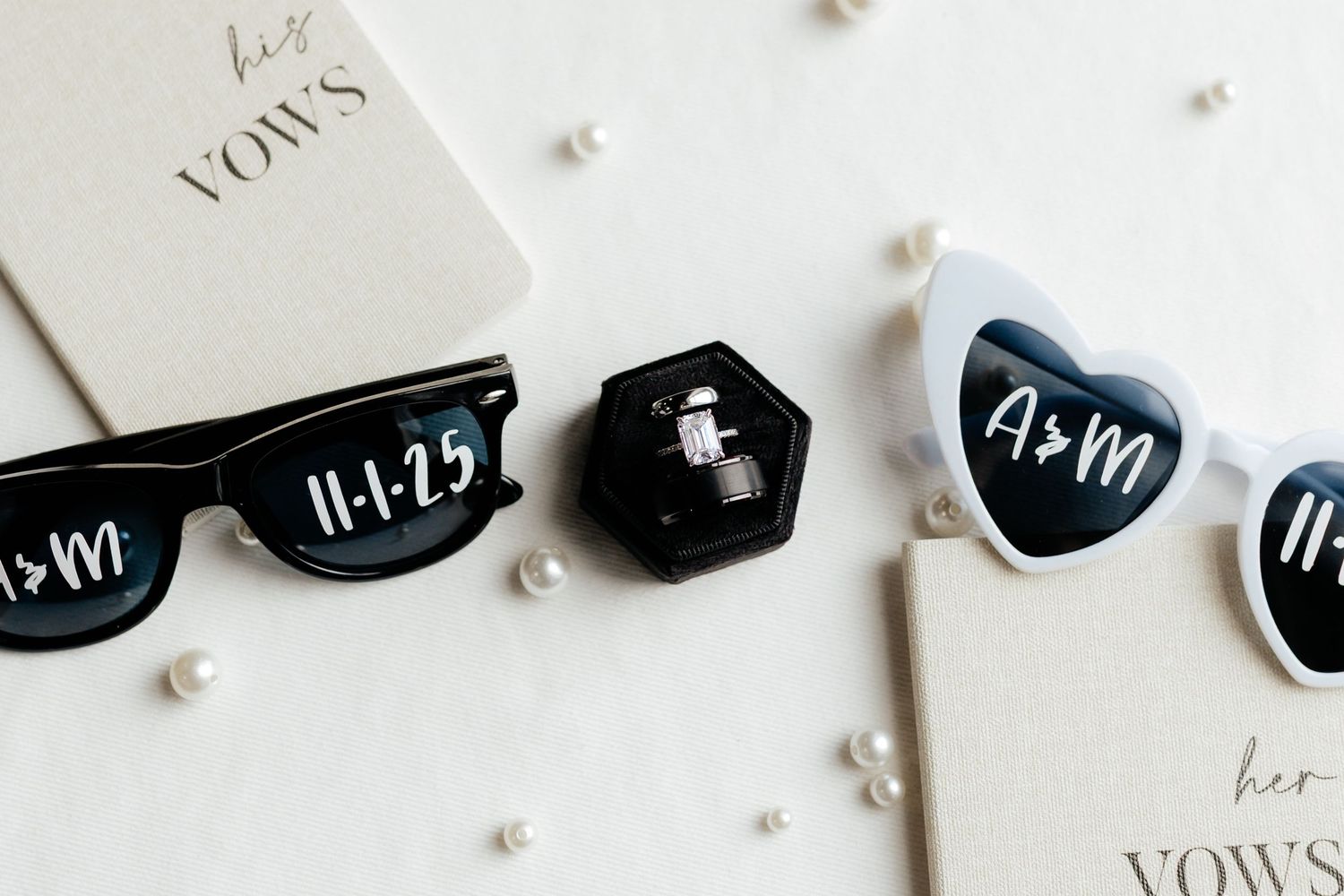 Wedding detail flat lay with custom date sunglasses, vow books, pearl accents, and engagement ring box.