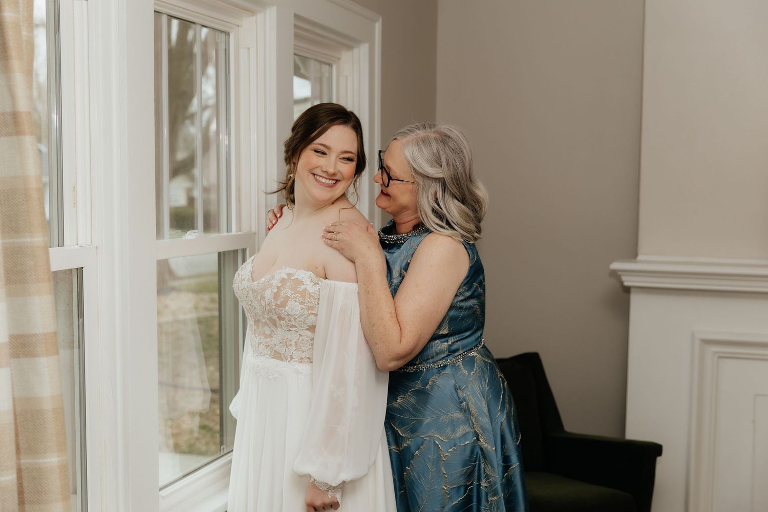 Bride in lace wedding gown sharing a joyful moment with her mother near a bright window.