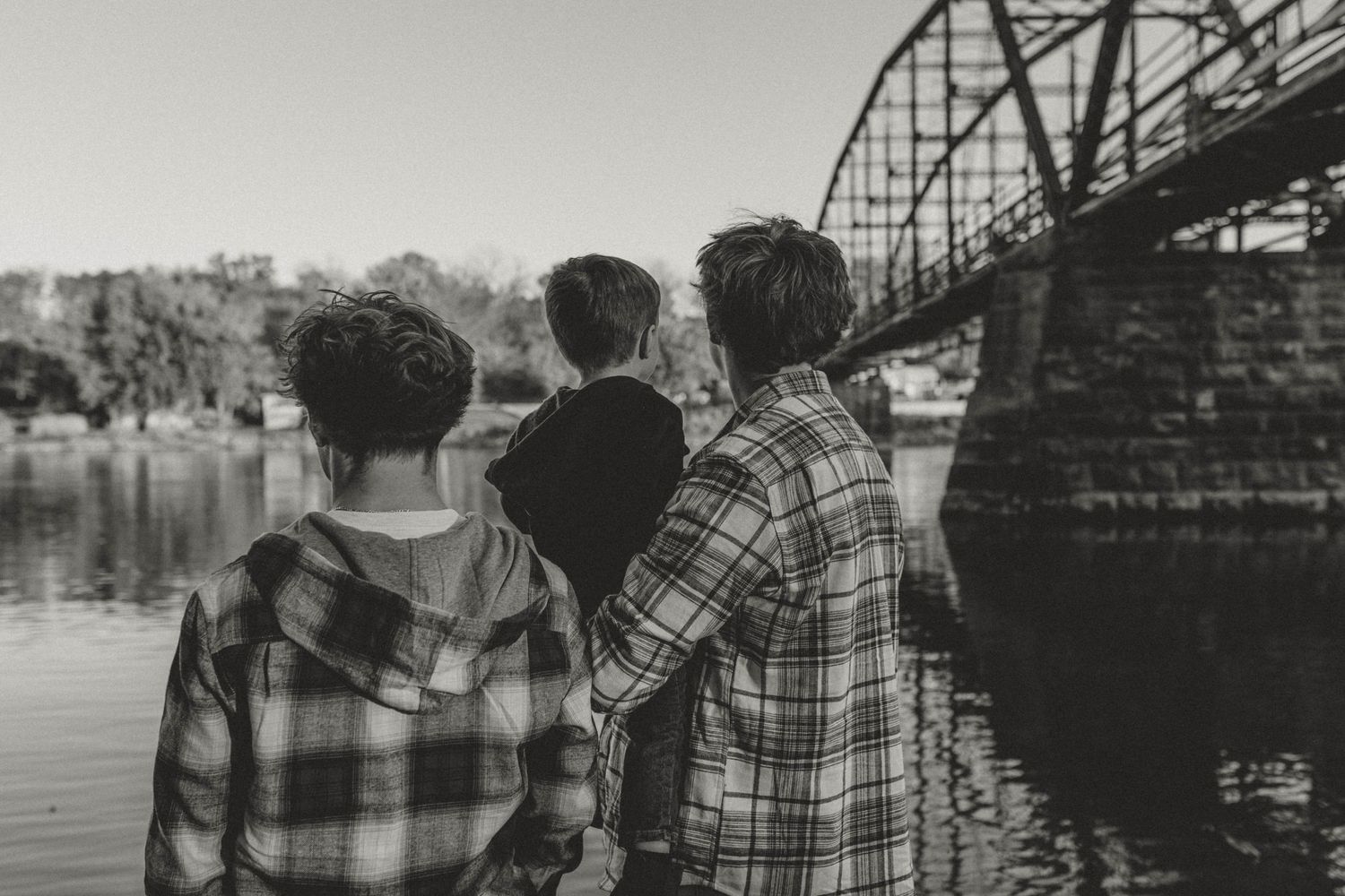 Three teens in plaid shirts stand by a river with an iron bridge in black and white photo.