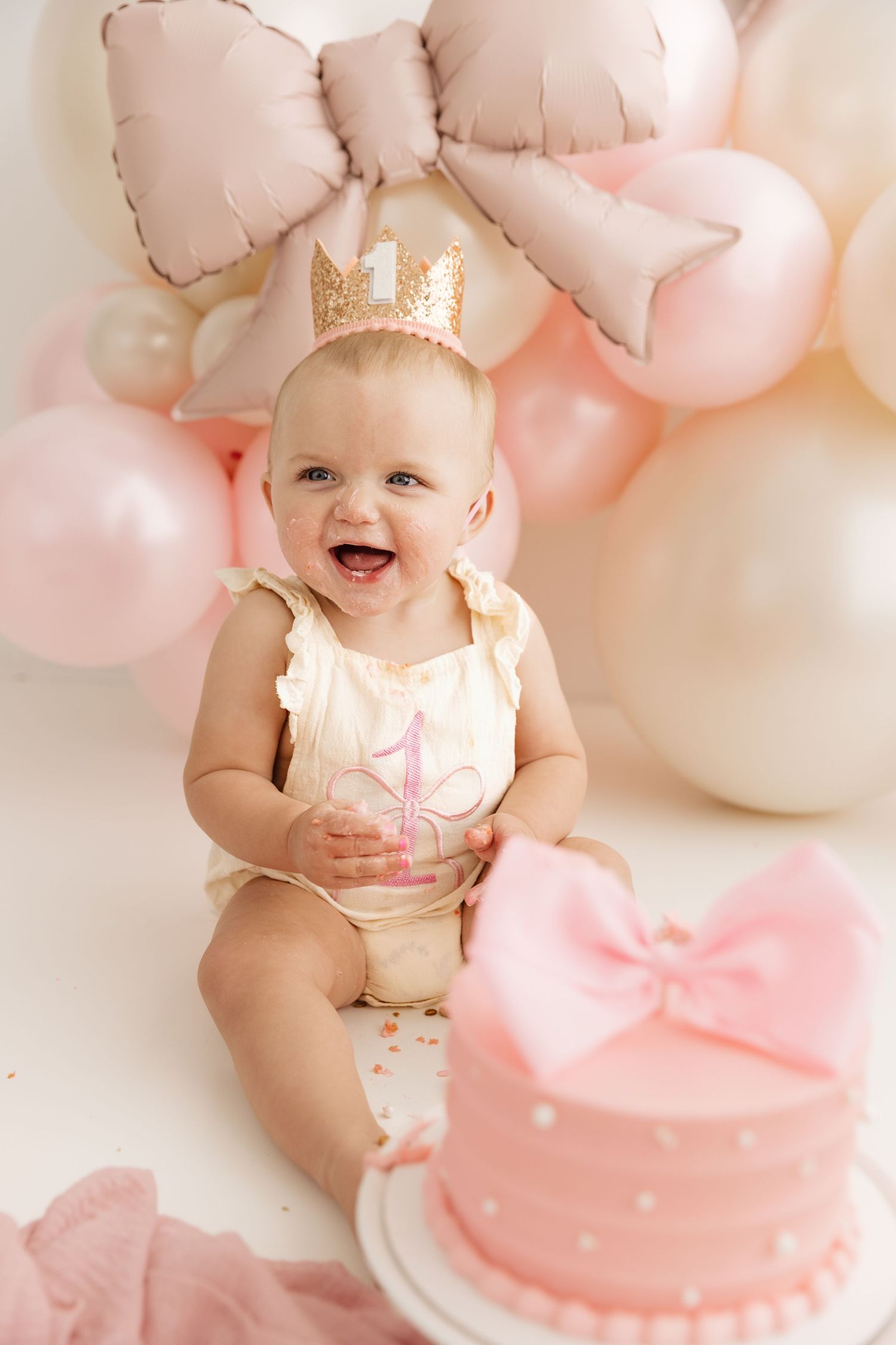 Laughing baby girl in crown and cream romper celebrating first birthday with pink bow cake and balloons.