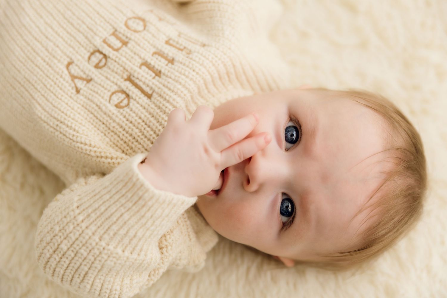 Newborn baby in cream 'little one' knit sweater lying on fluffy white blanket, chewing on fingers.