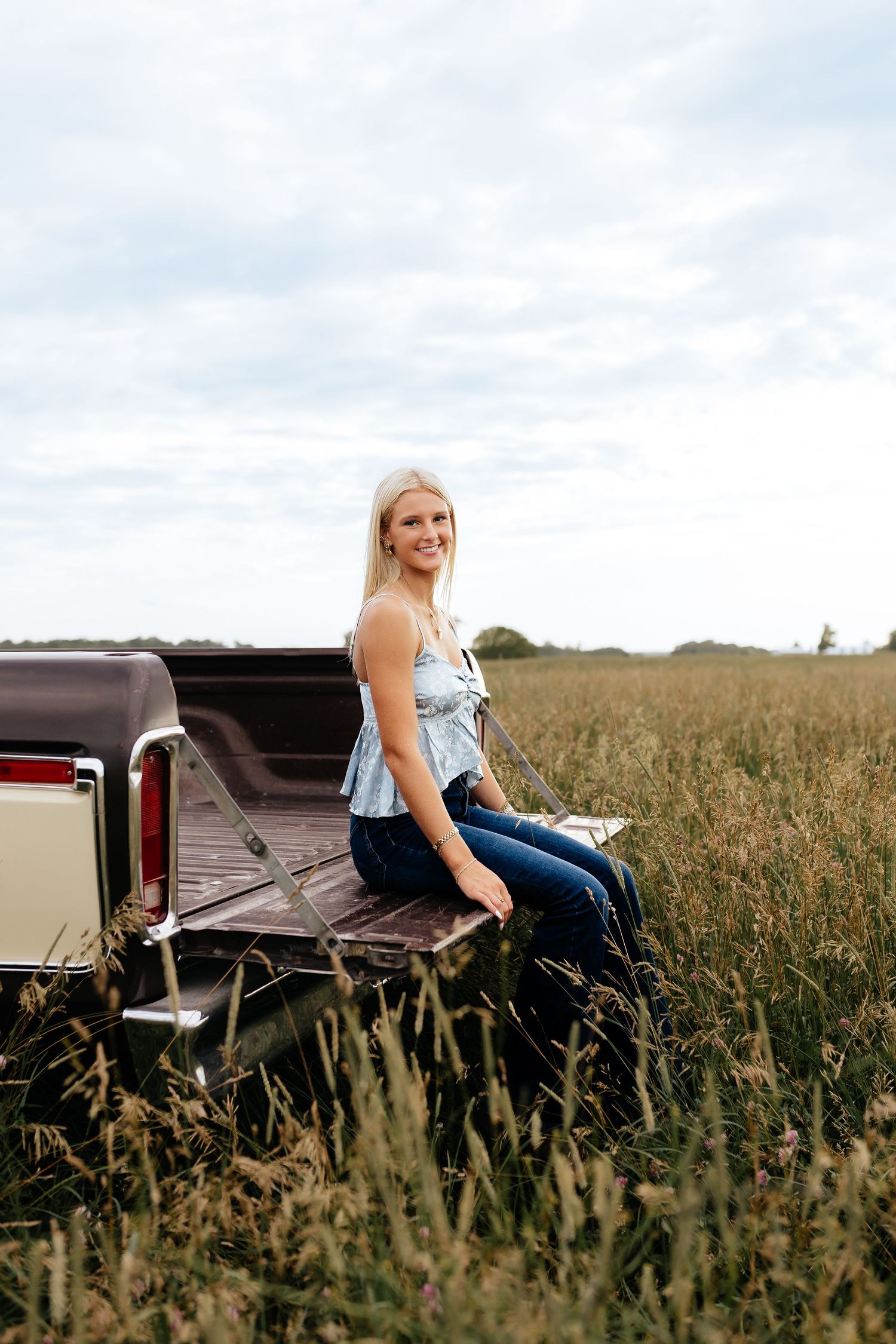 A blonde girl sits on the tailgate of a vintage pickup truck in a golden field under cloudy skies.