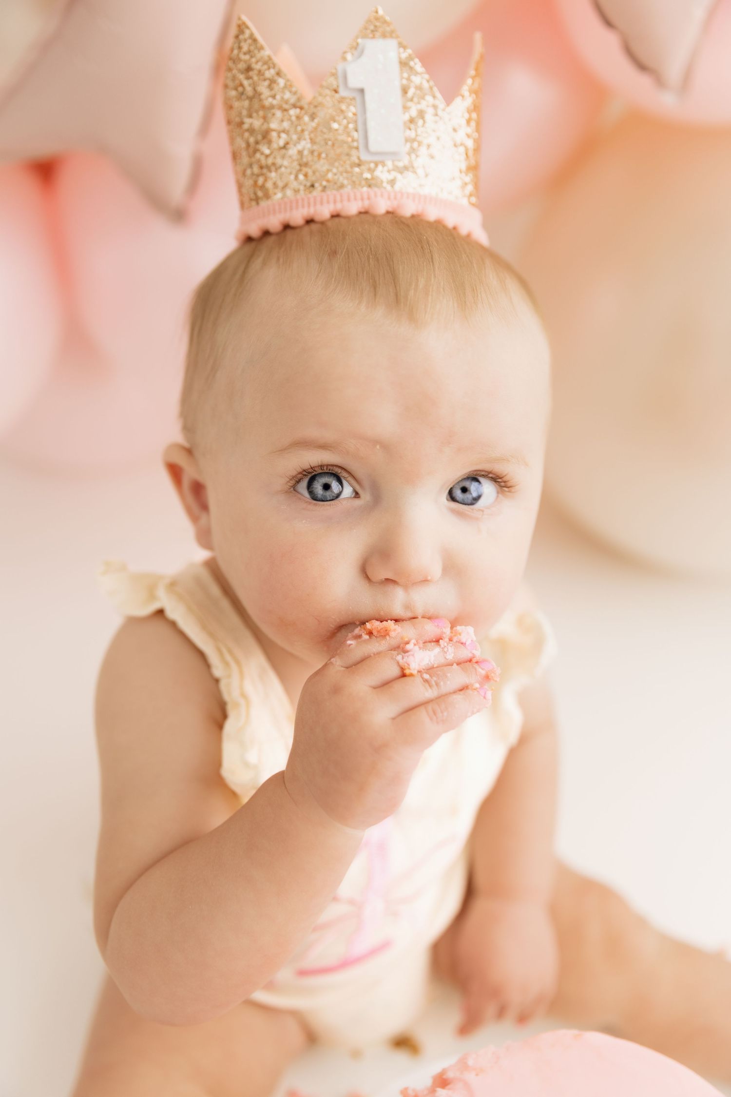 Baby girl in cream outfit wearing gold number one crown eating birthday cake at first birthday celebration.
