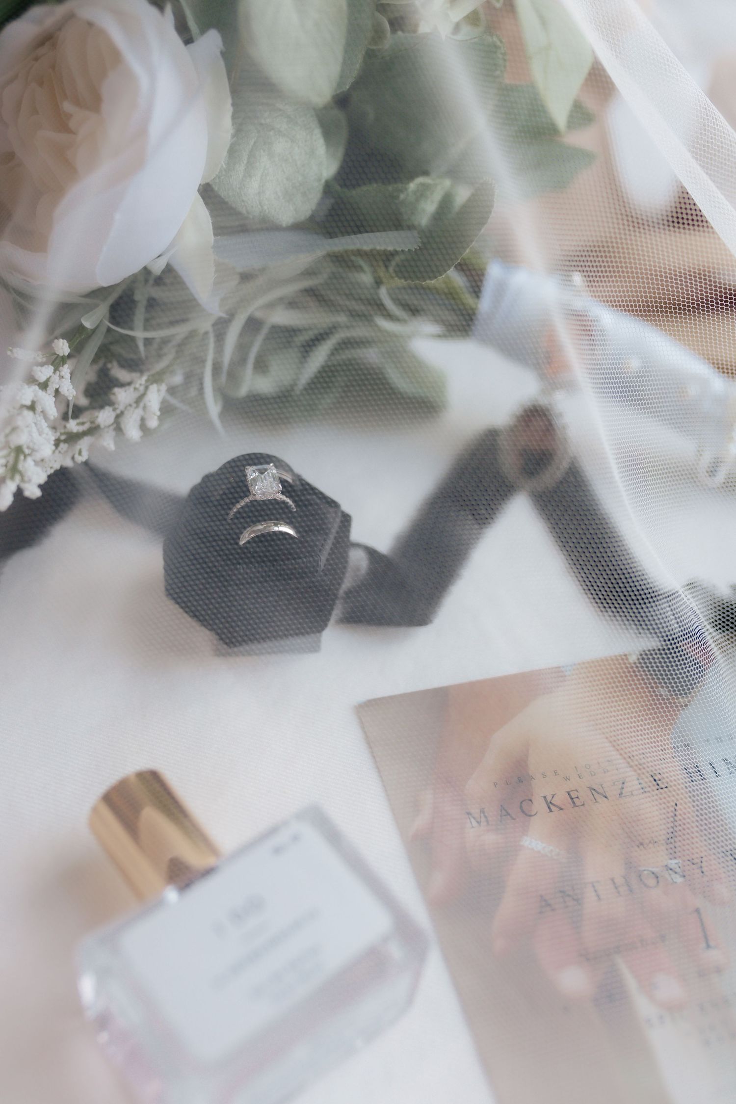 Bridal flat lay with diamond engagement ring in black velvet box, perfume, flowers, and wedding invitation.