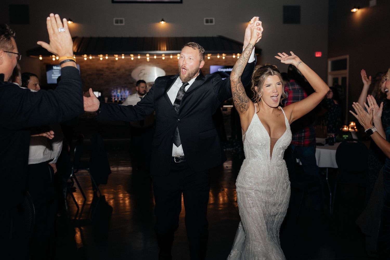 Bride in lace gown and groom in tuxedo celebrate joyfully on crowded wedding dance floor.