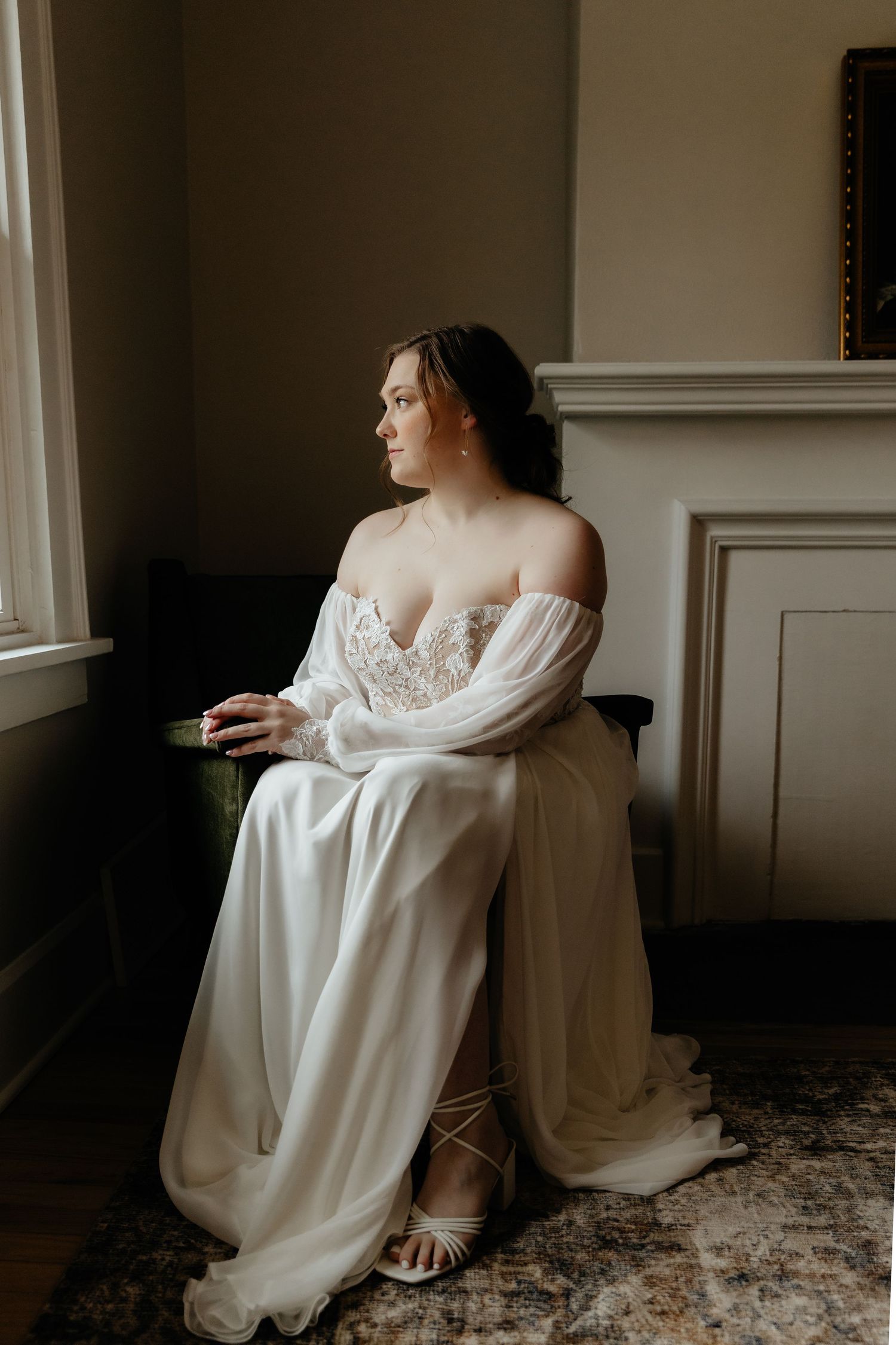 Elegant bride in off-shoulder lace wedding gown sitting by window in soft natural light.