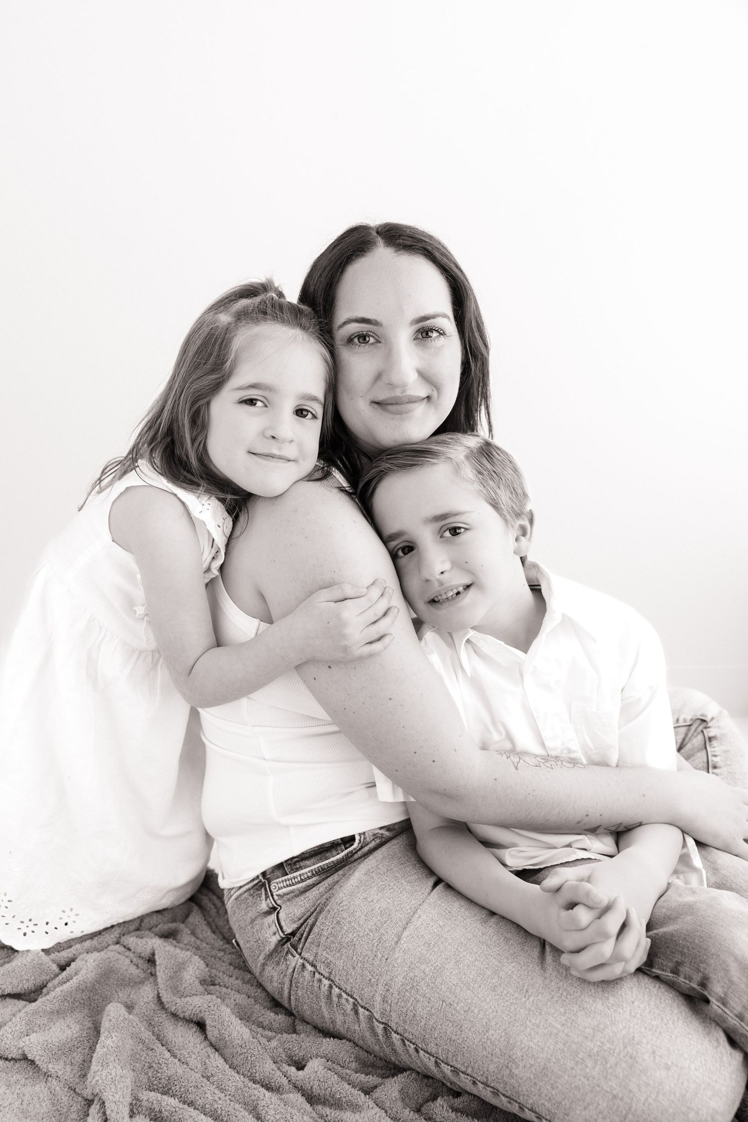 Black and white photo of a smiling mother hugging her two young children on a blanket.