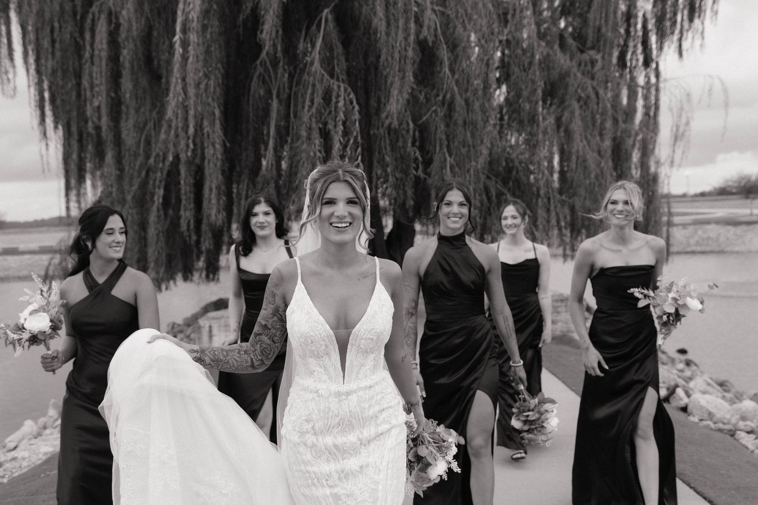 Black and white photo of a bride in a lace gown walking with her bridesmaids in black dresses.