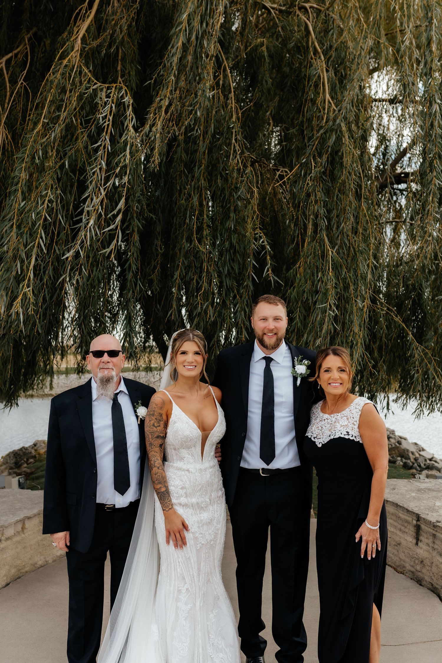 Bride and groom smile with two family members under weeping willow at outdoor wedding venue.