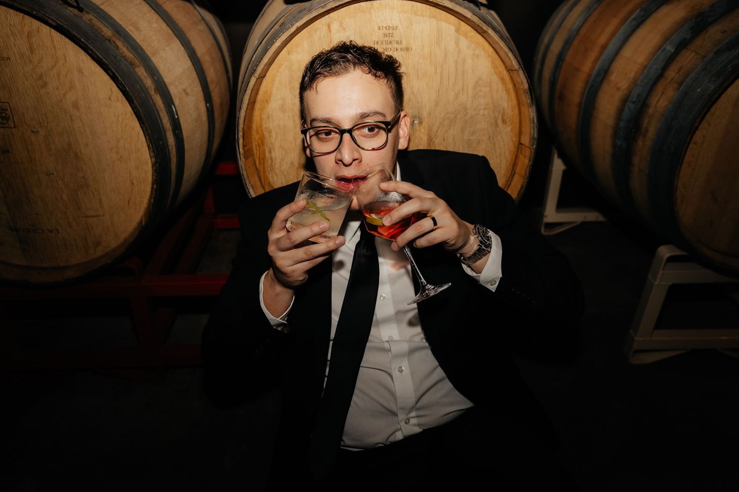 Person in black tuxedo holds two cocktail glasses while seated in front of wooden wine barrels at winery.
