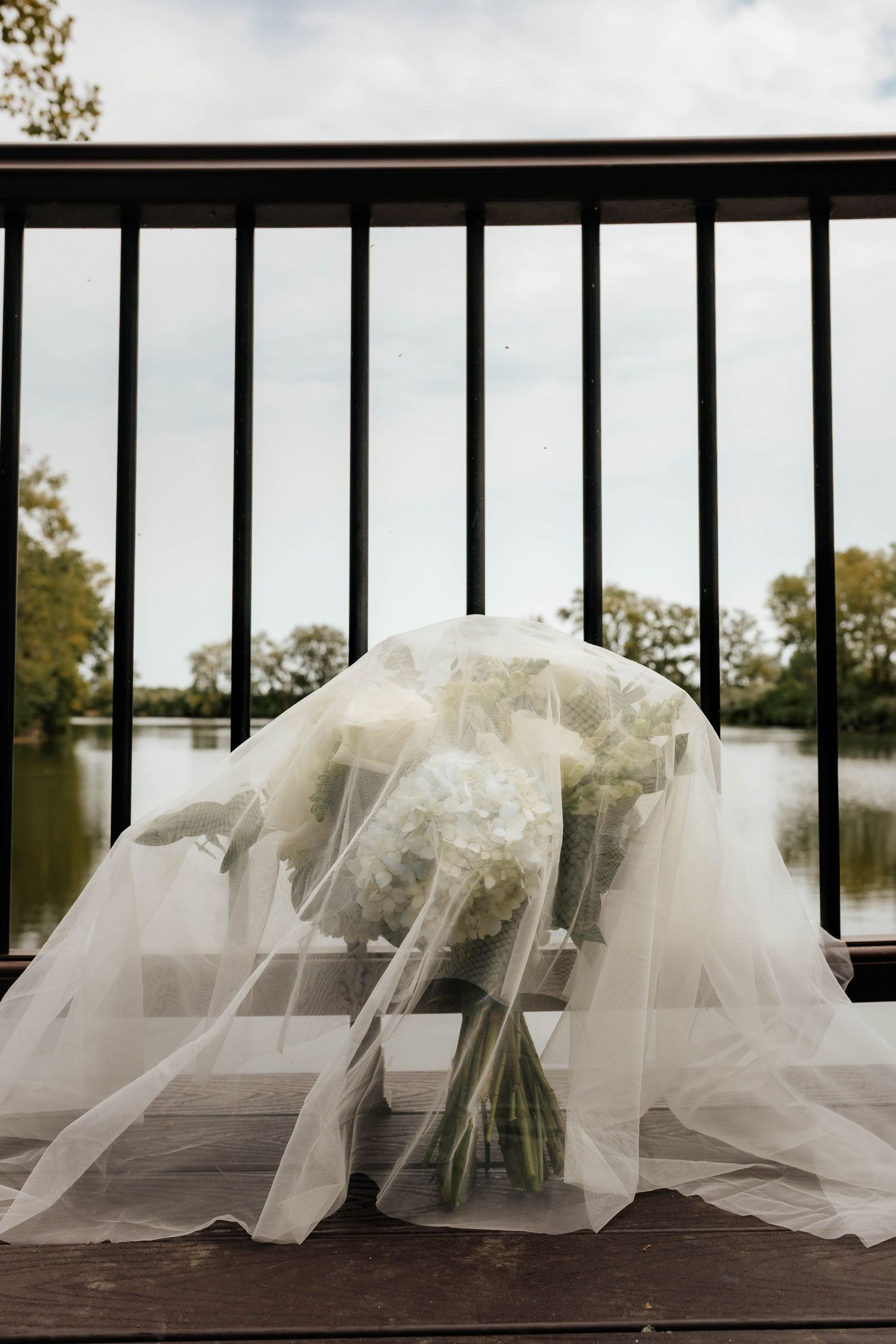 Bridal bouquet of white hydrangeas draped with a flowing veil on a wooden deck overlooking a serene lake.