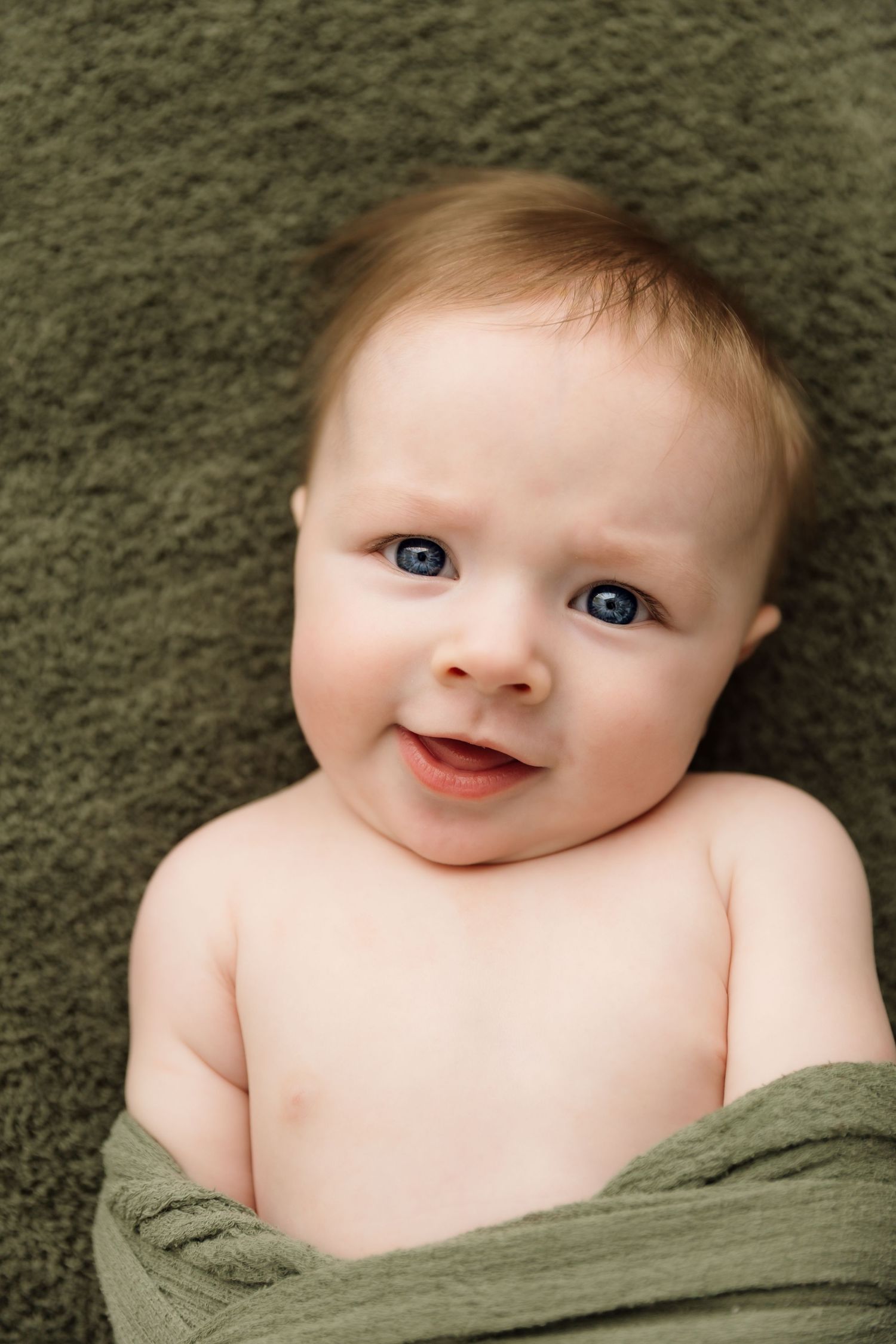 Adorable blue-eyed baby wrapped in green cloth smiling on a green blanket.