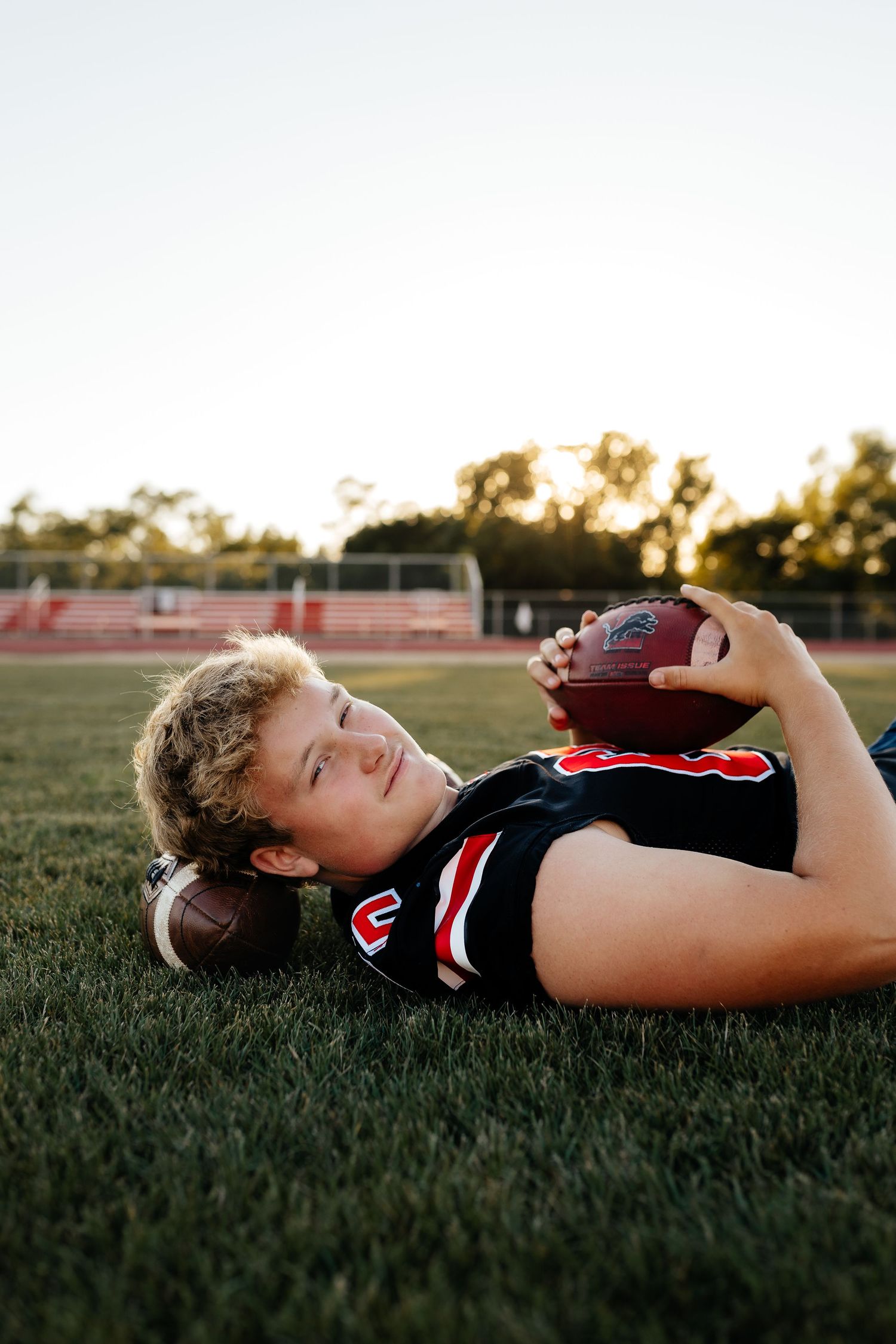 Young male football player in black jersey lying on grass holding a football at sunset.