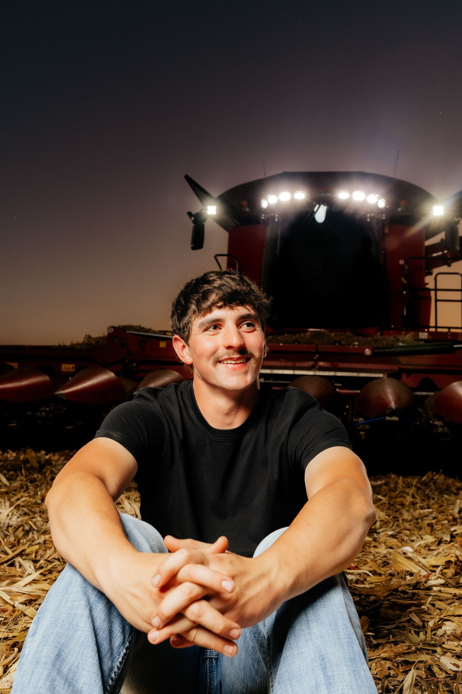 Smiling young man sits in harvested field at night with lit farm combine in background.
