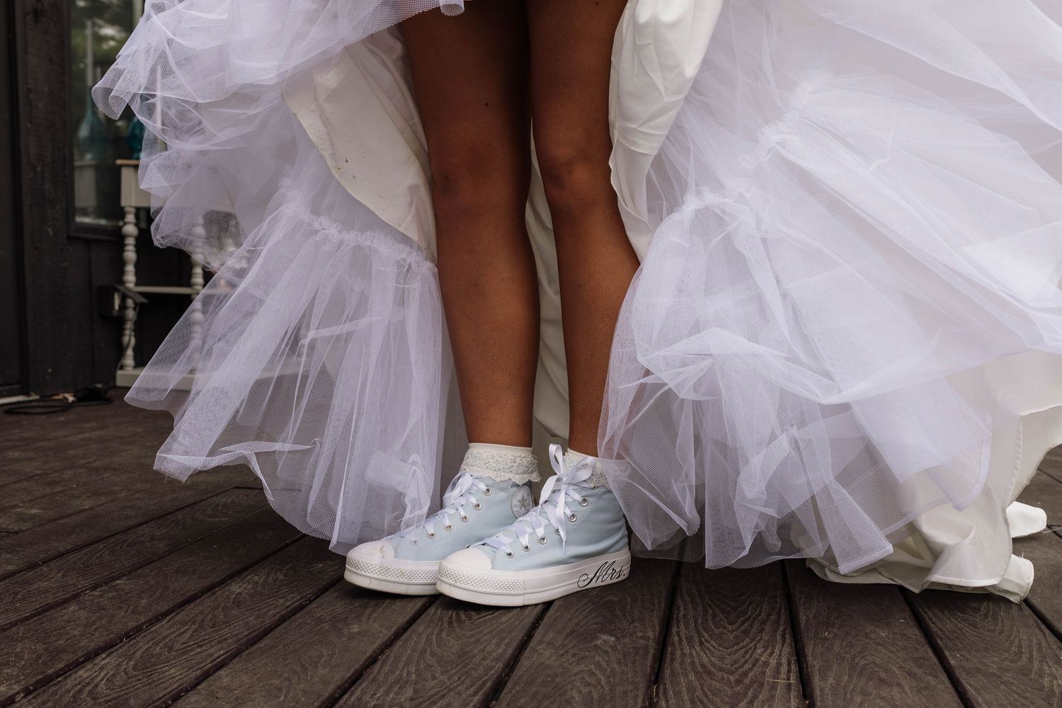 Bride wearing white tulle wedding gown paired with casual light blue Converse sneakers on a wooden deck.