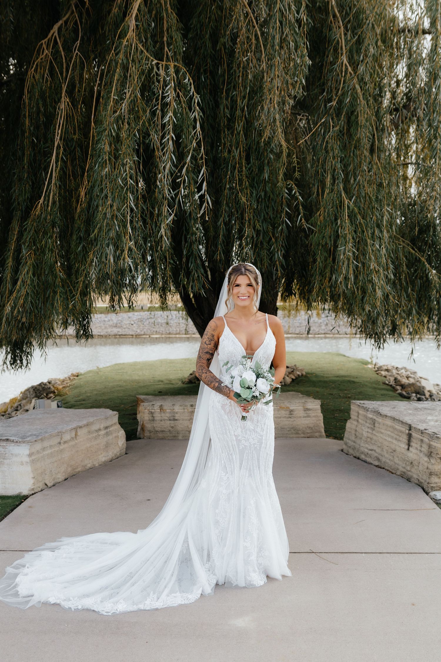 Bride in backless lace mermaid gown holds white bouquet while posing under weeping willow tree.