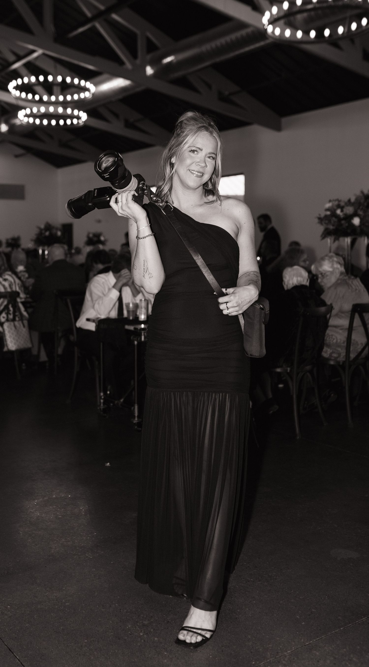 Woman in black one-shoulder gown holding camera at elegant indoor event venue with chandeliers.