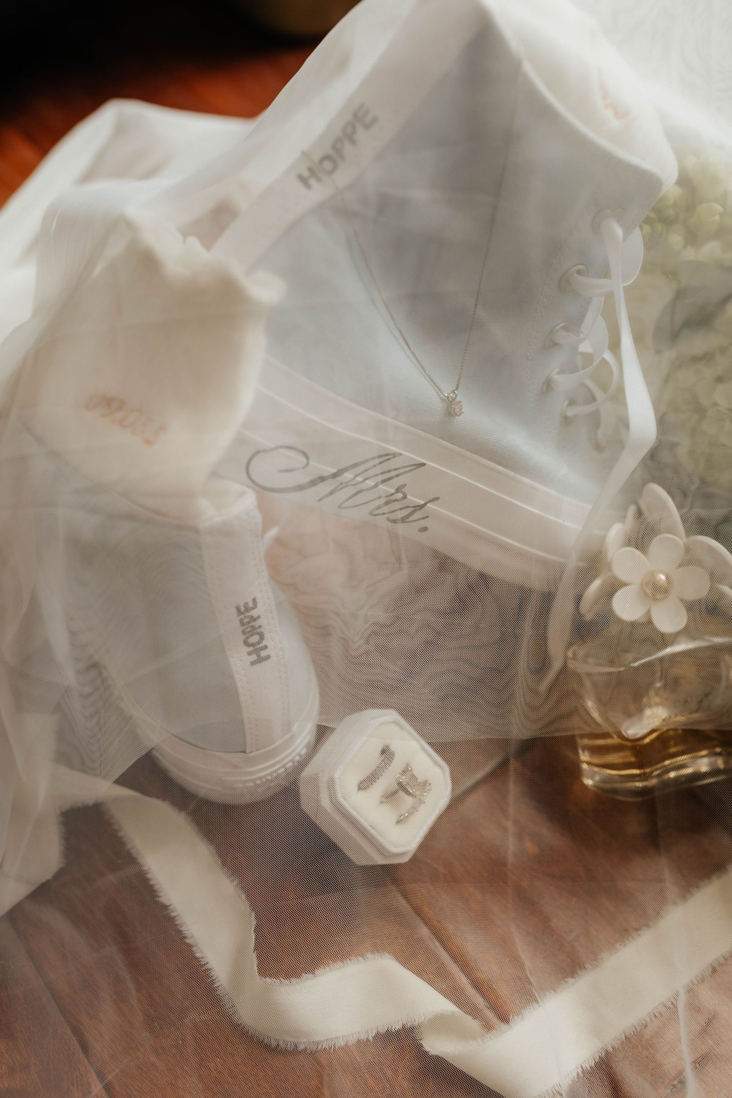 Bridal flat lay with white Converse Mrs. sneakers, veil, ring box, and perfume on wood floor.