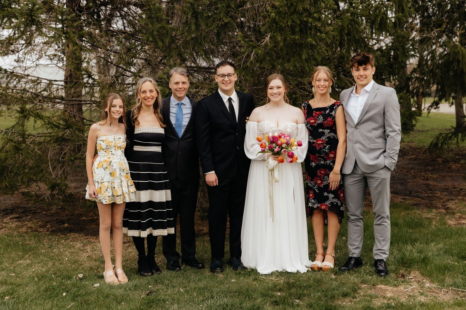 Wedding couple poses with family of five outdoors on green lawn near trees.