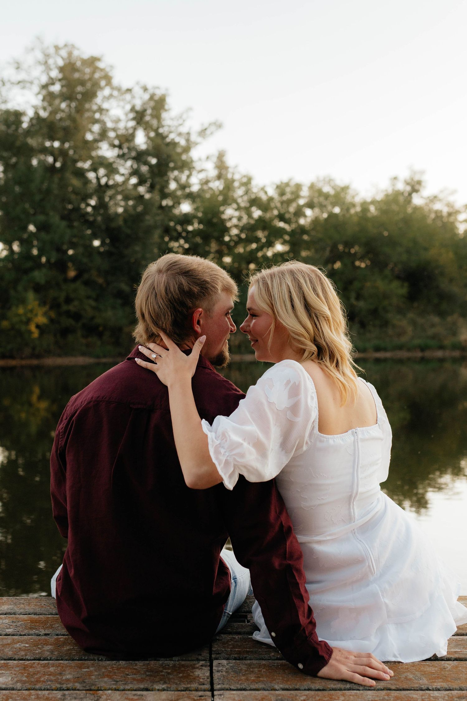 Couple sitting on wooden dock facing each other about to kiss with lake and trees behind them.