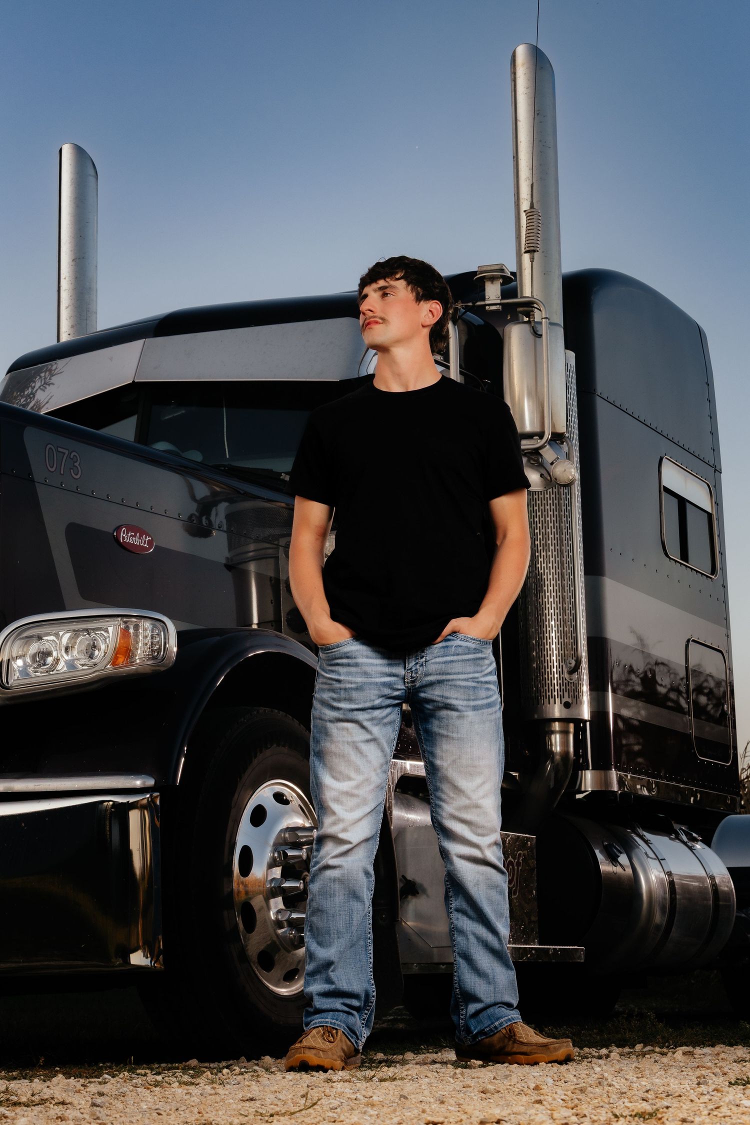 Young man in black t-shirt and jeans leaning against a large black Peterbilt semi truck outdoors.