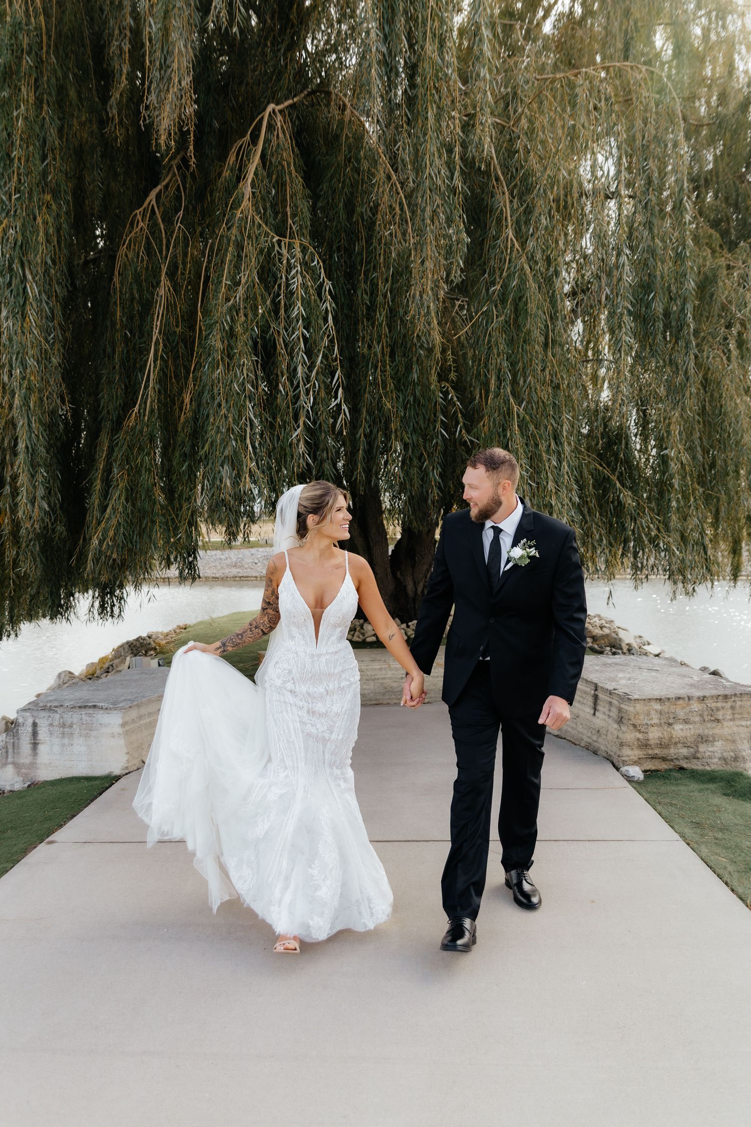 Bride and groom walk hand in hand under weeping willow, bride's lace train flowing behind her.