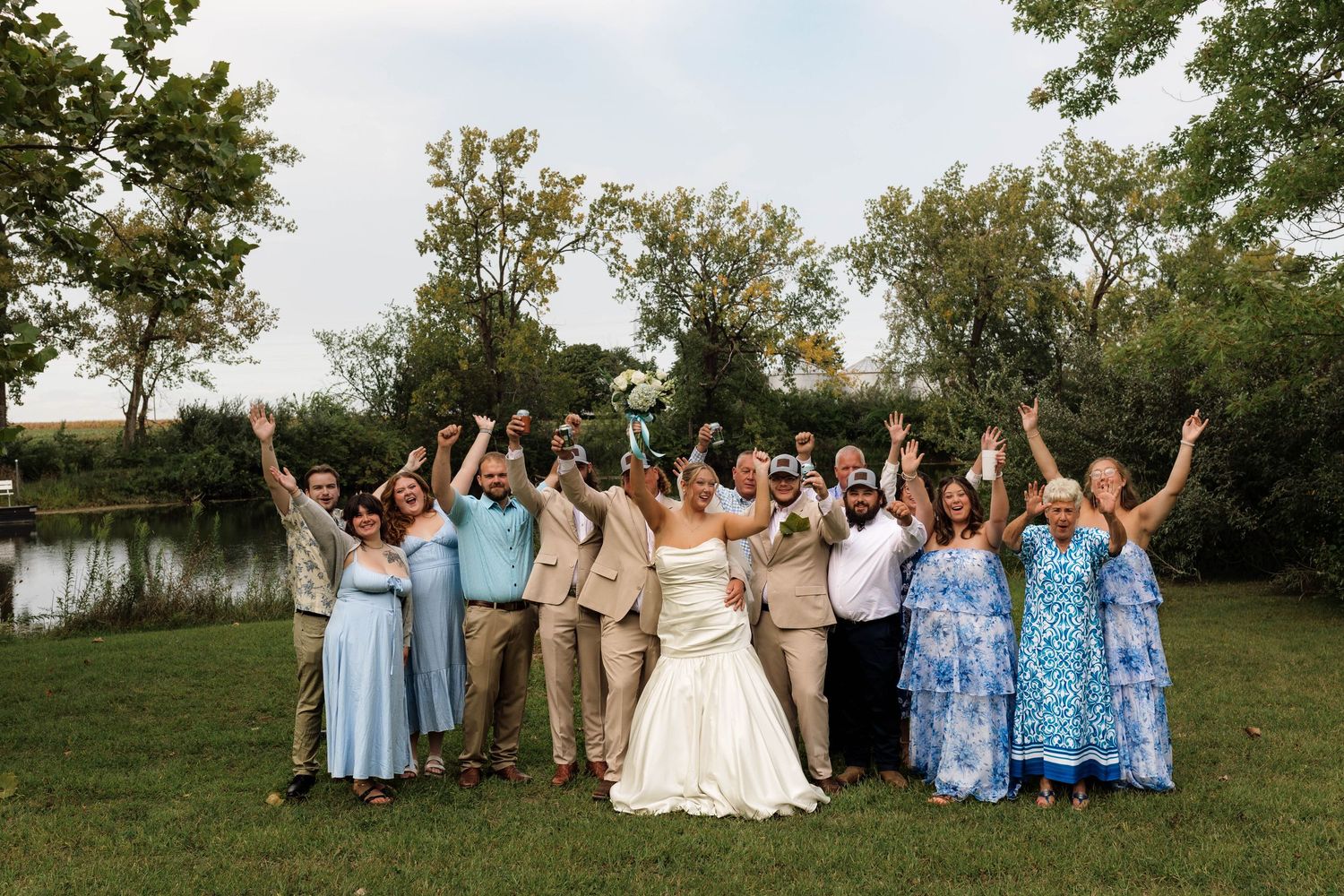 Large wedding party celebrates with raised arms on a green lawn beside a beautiful tree-lined pond.