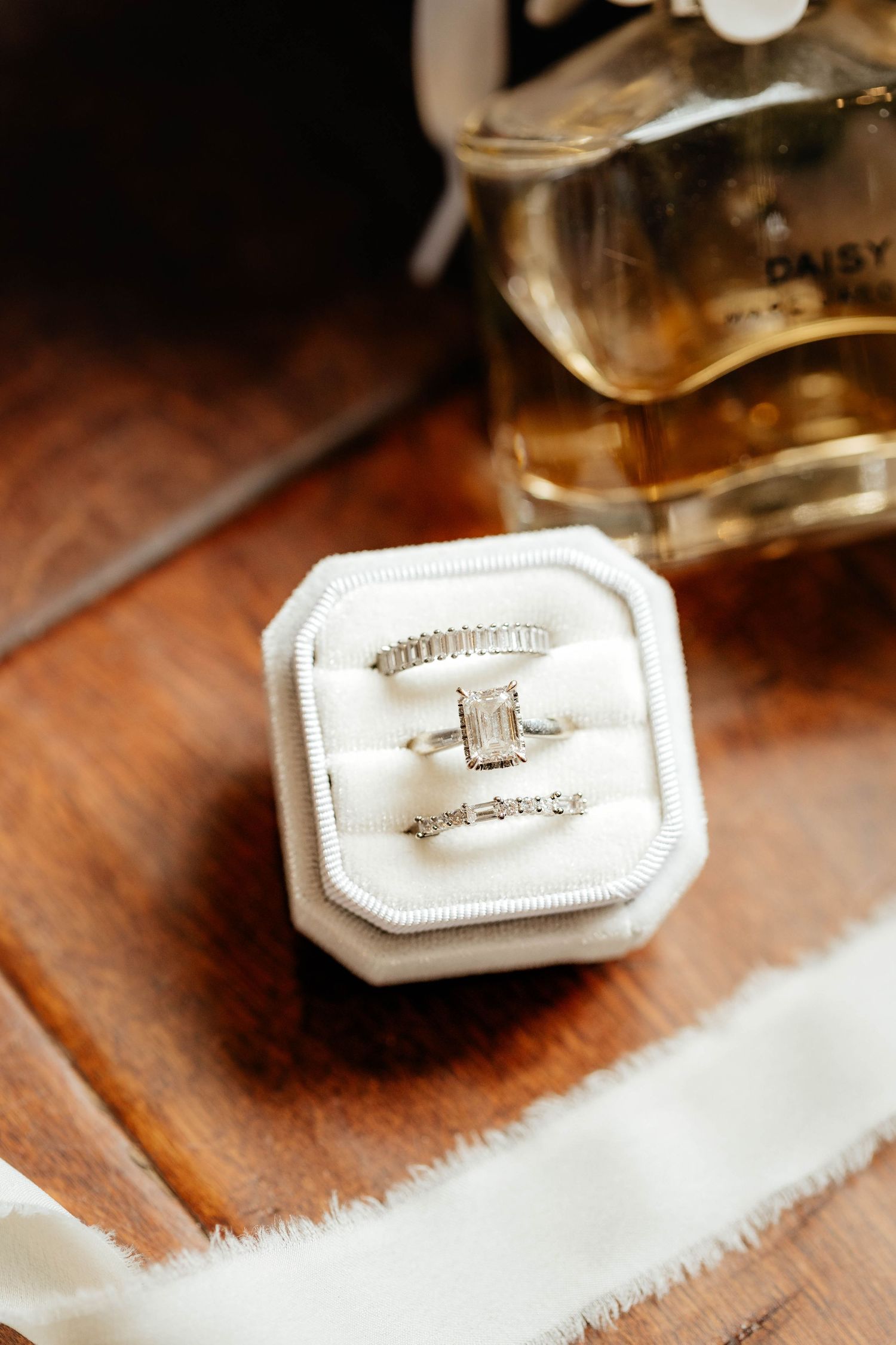 Elegant emerald-cut diamond engagement ring with wedding bands in white velvet box beside Daisy perfume.