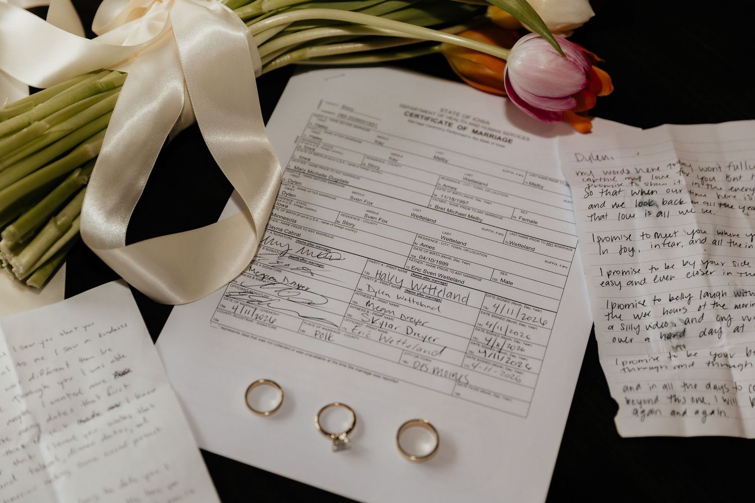 Wedding certificate with gold rings, handwritten vows, and floral bouquet on dark surface.