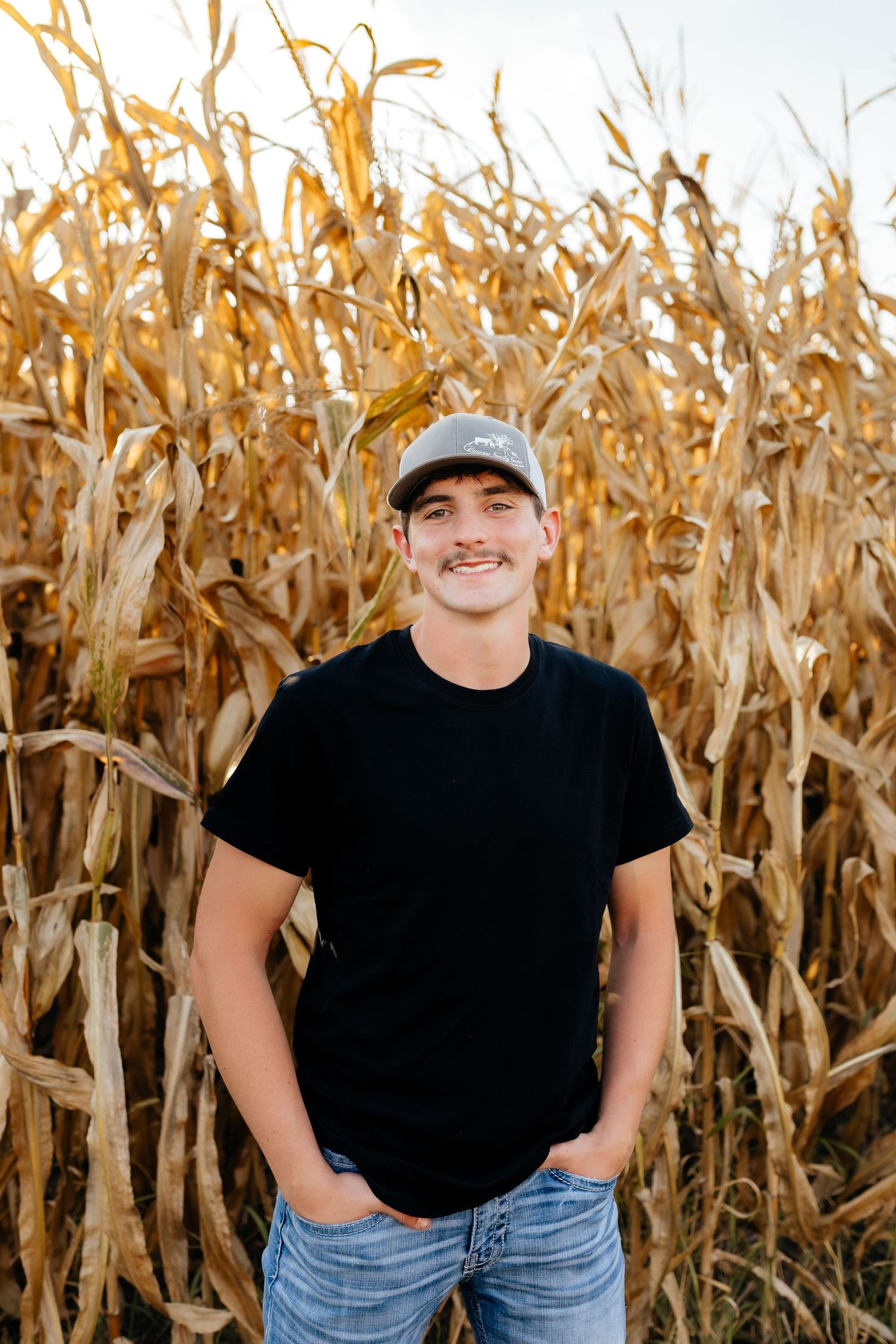 Smiling young man with mustache in black tee and cap standing before golden dried corn stalks.