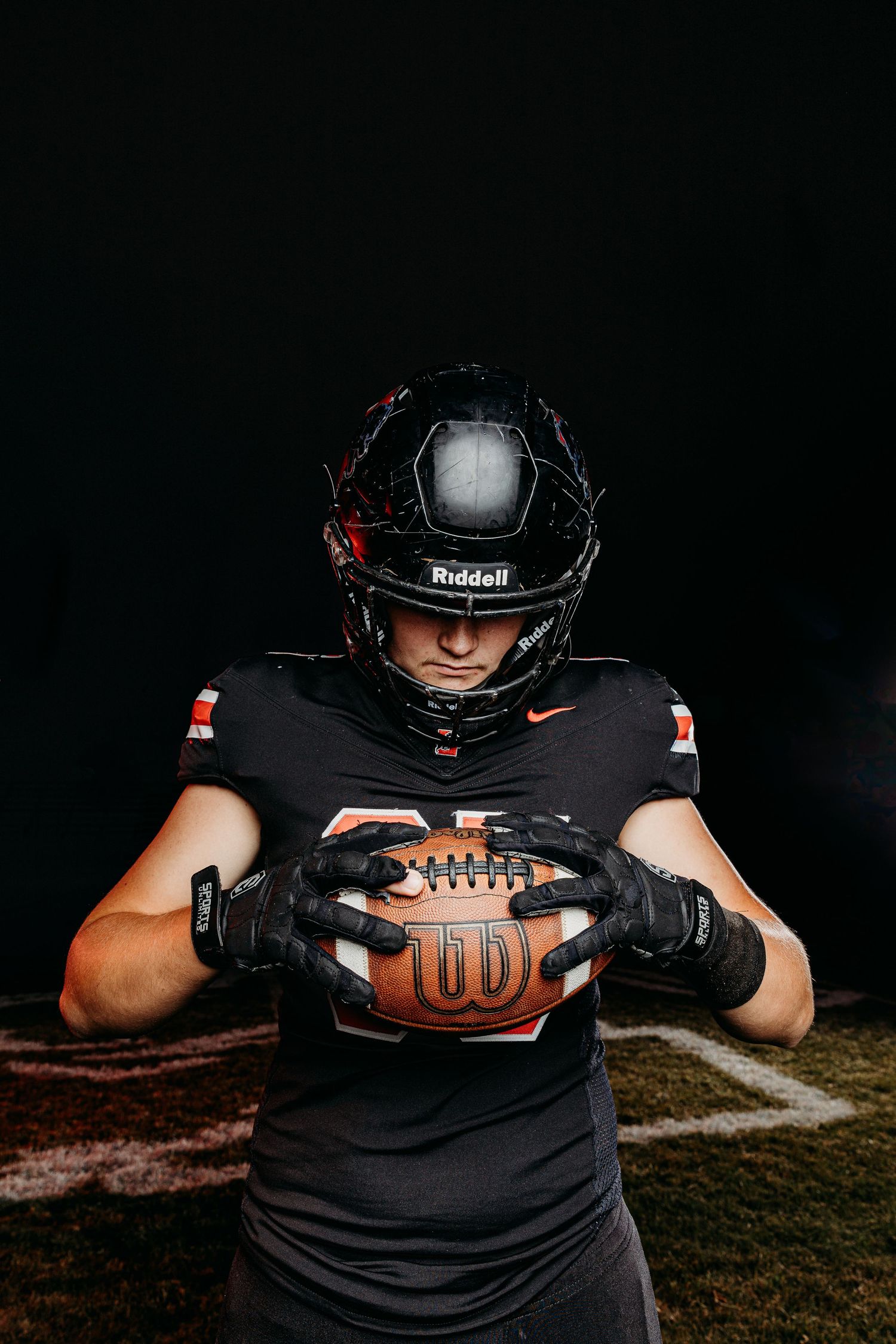 Football player in black Riddell helmet and gloves holding a Wilson football on a dark field.