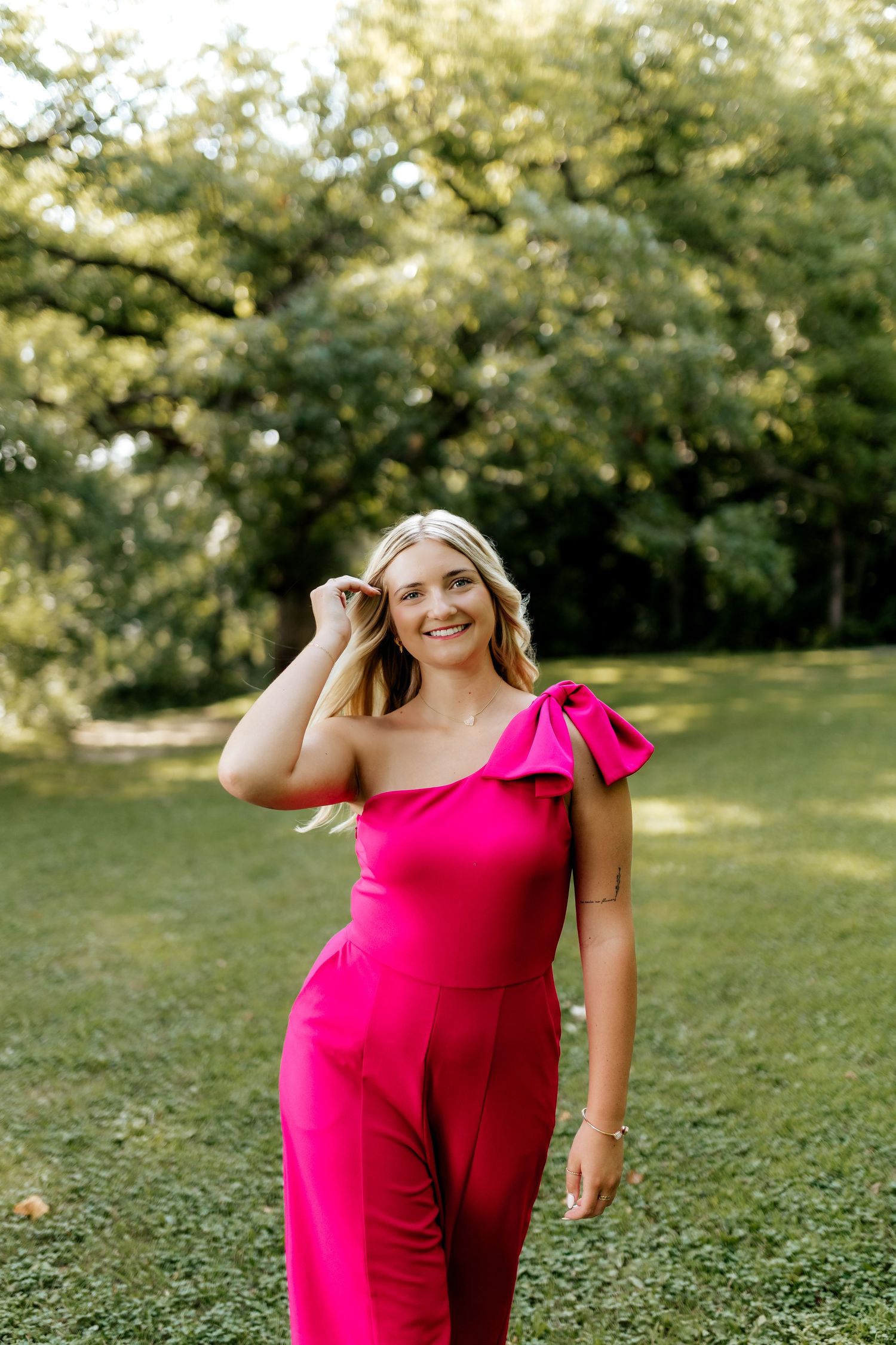 Smiling blonde woman in a hot pink one-shoulder ruffled jumpsuit posing in a lush green park.