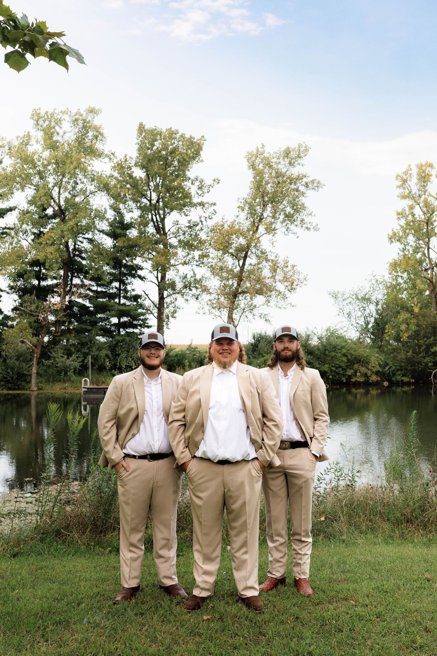 Three groomsmen in matching tan suits and baseball caps stand confidently by a peaceful lakeside setting.