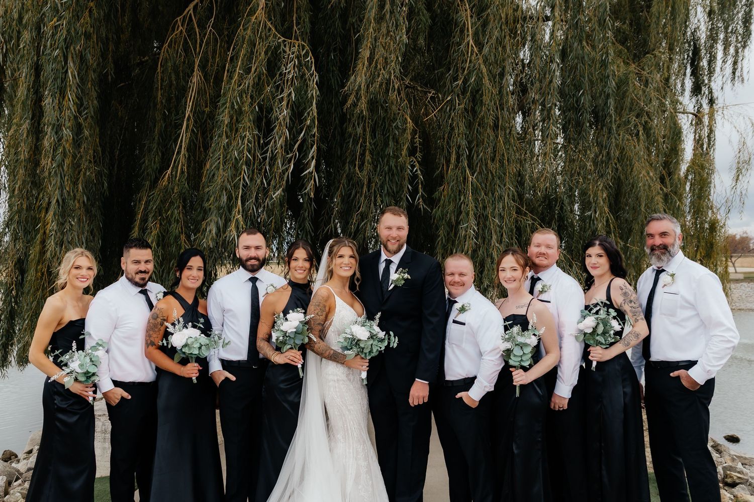 Wedding party in black attire posing with bride and groom near weeping willow tree.
