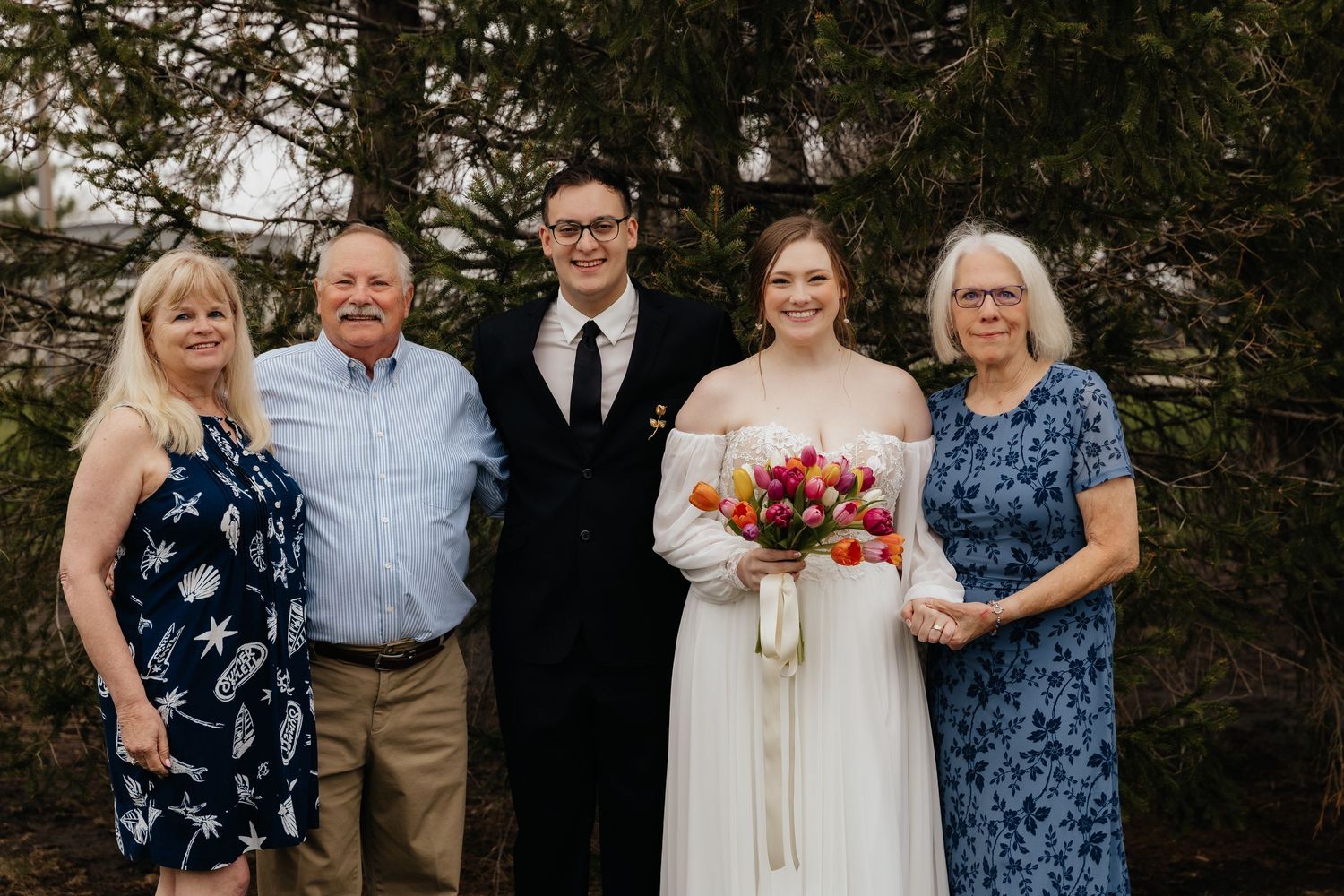 Wedding couple poses with family of four outdoors near evergreen trees.