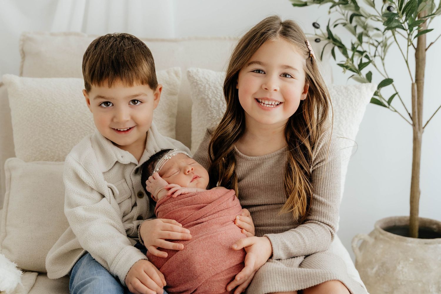 Boy and girl siblings sit together smiling while holding their pink-swaddled newborn baby.