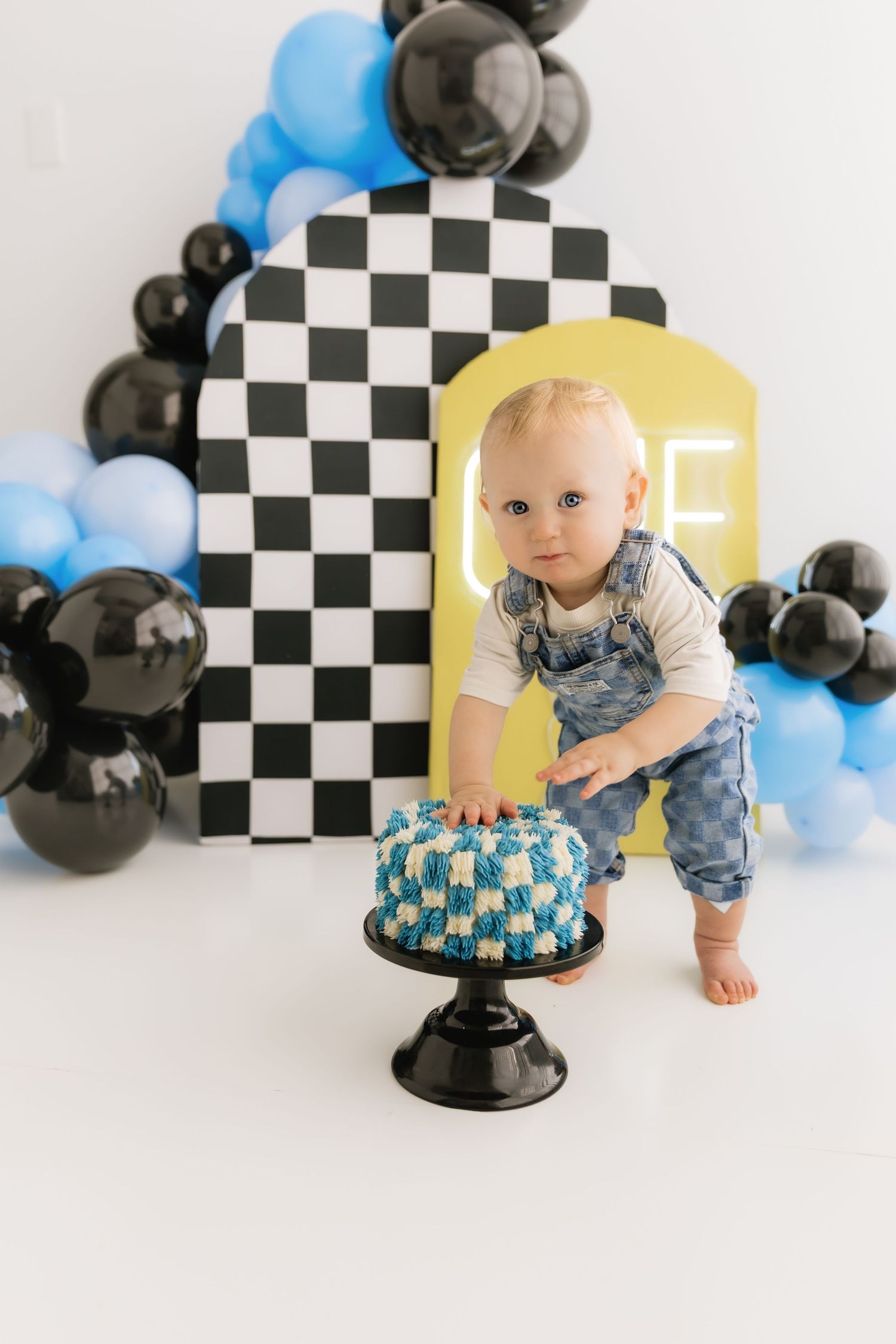 Baby boy in denim overalls reaching toward blue and white checkered smash cake at race car themed first birthday.