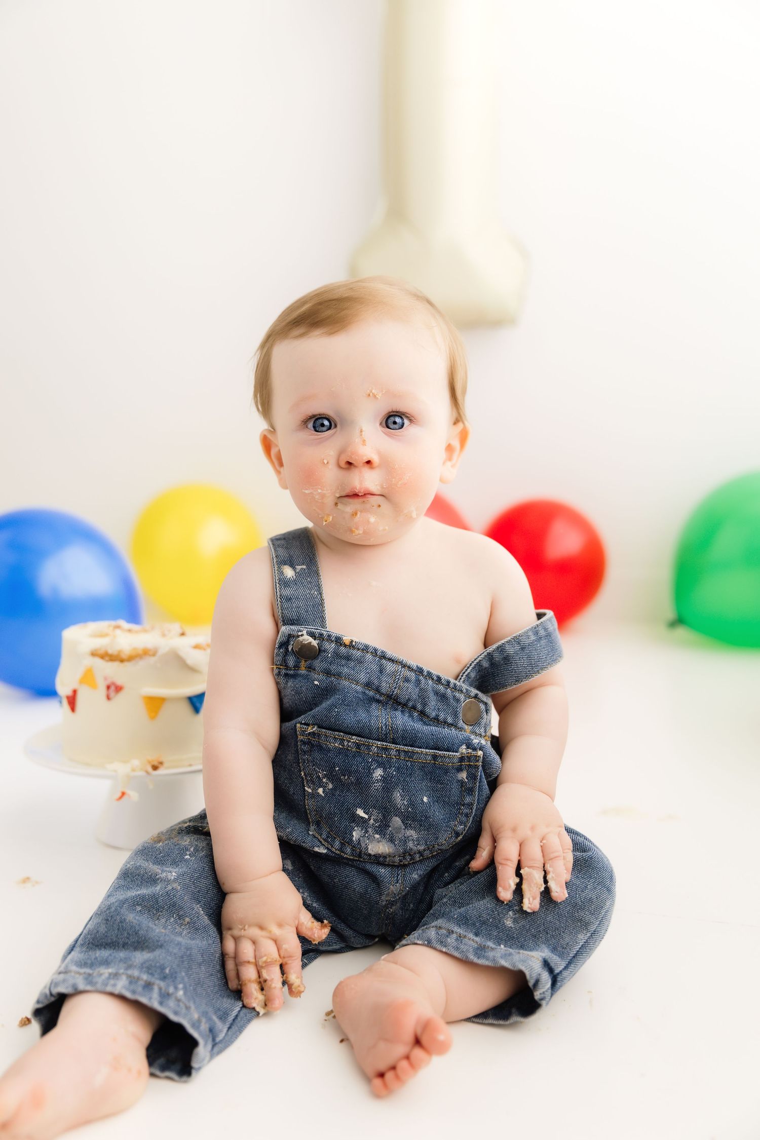 Adorable baby in denim overalls sitting beside smashed birthday cake with colorful balloons.