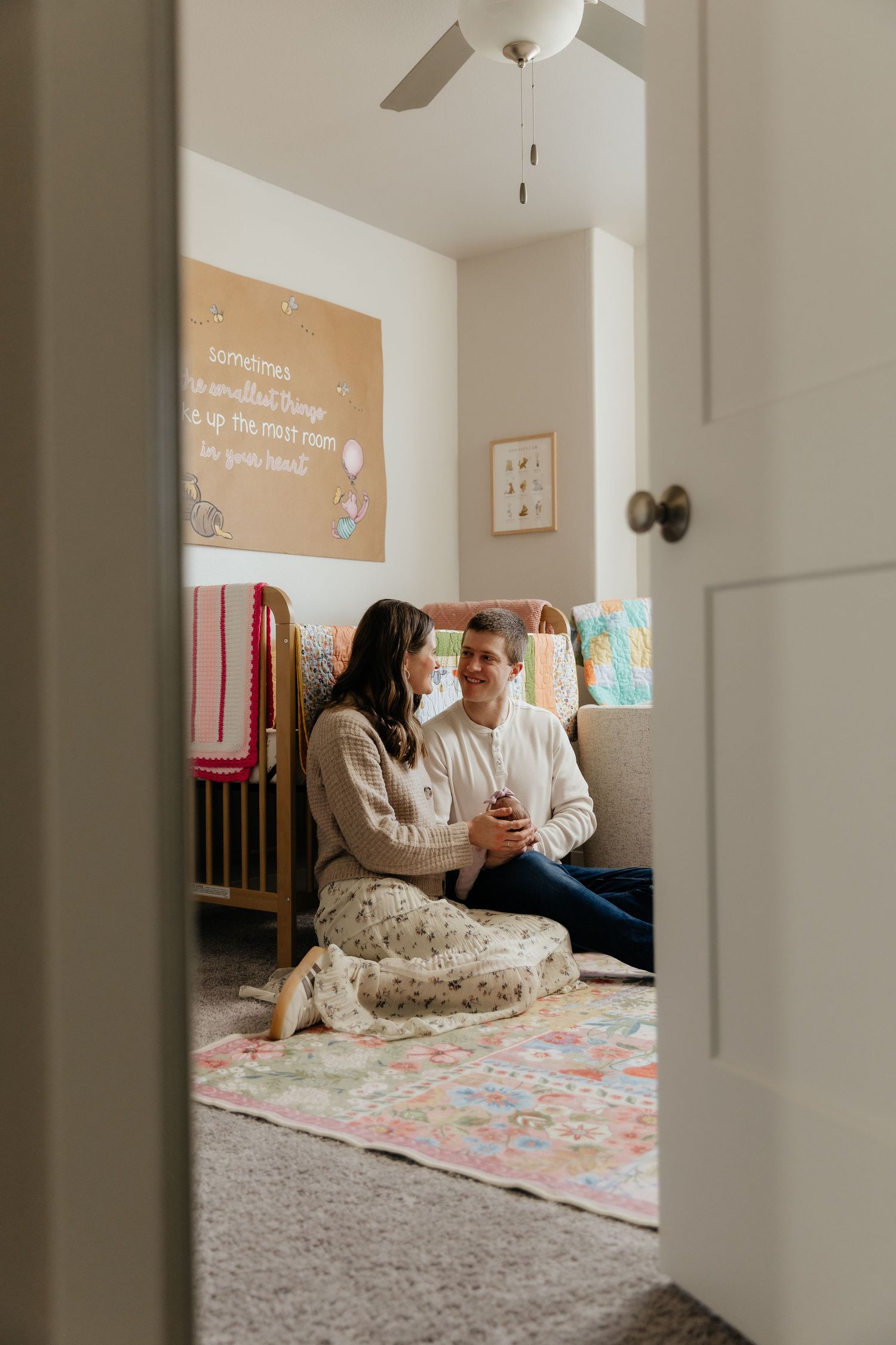 Couple sits together on nursery floor viewed through doorway with crib and decor.